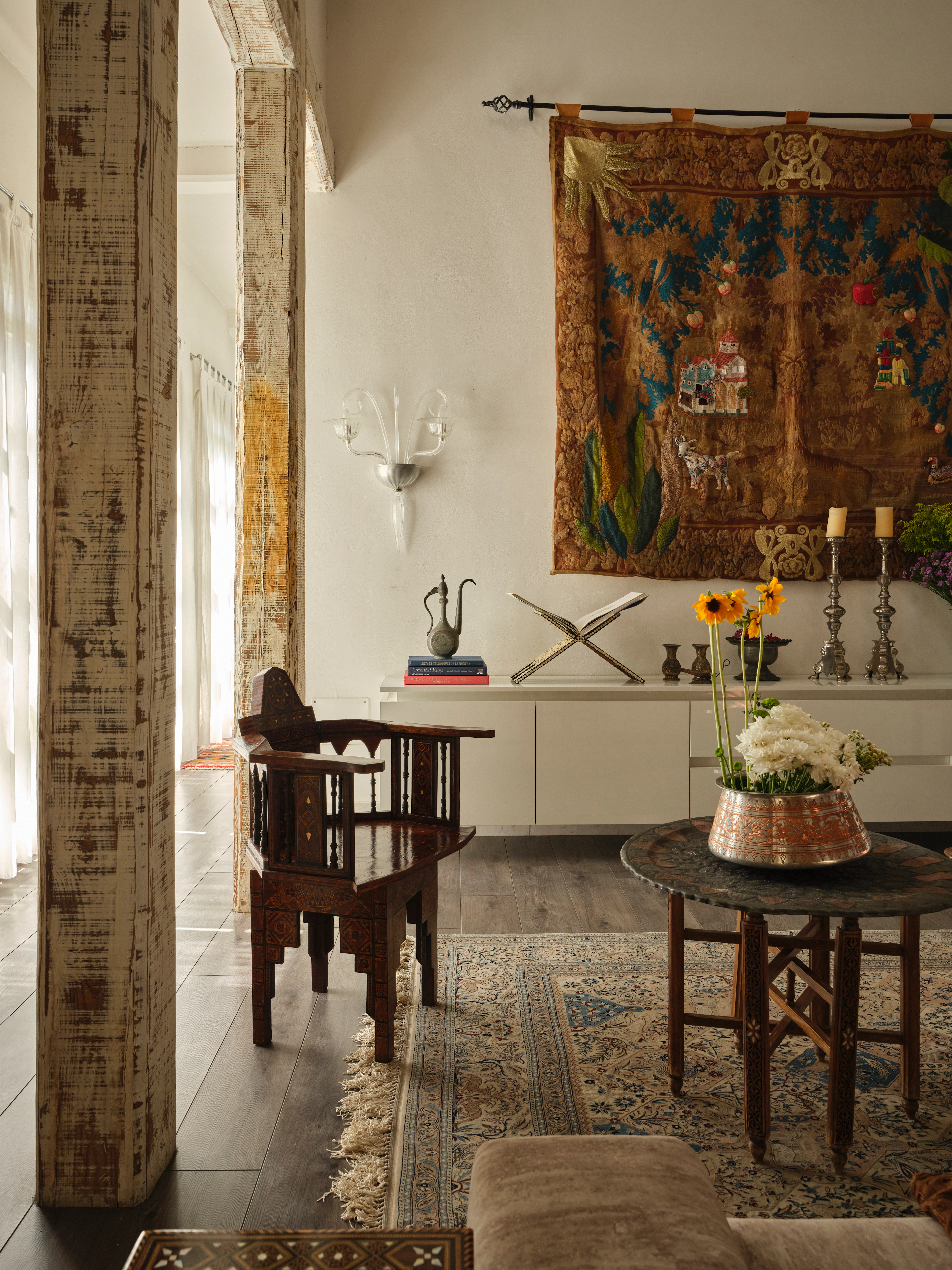 Detail of a living room with a colorful woven wall hanging, carved wooden chair, patterned rug, and flower arrangement.