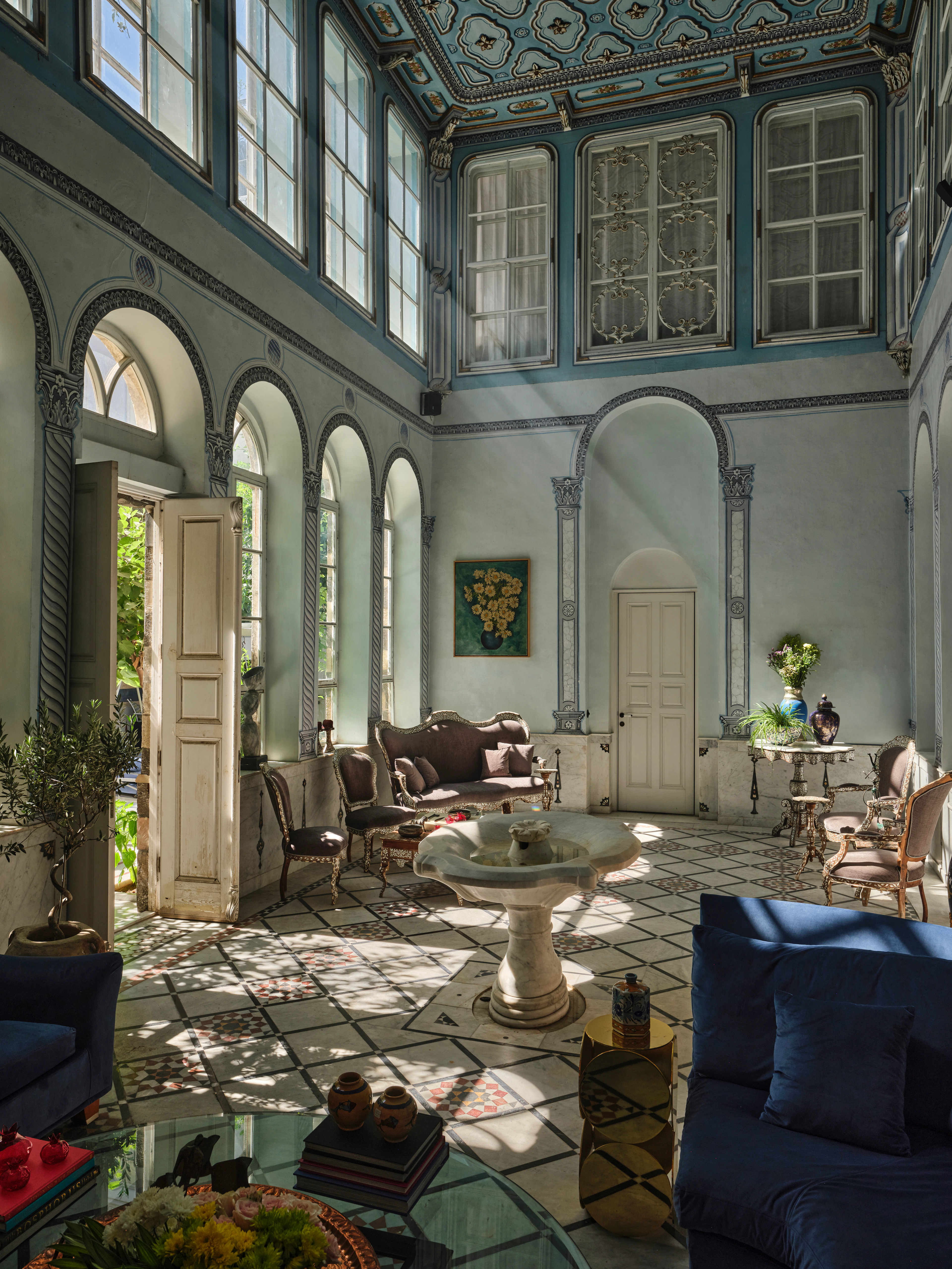 Sunlit blue and white salon with a painted ceiling, arched windows, patterned floor, and blue armchairs.