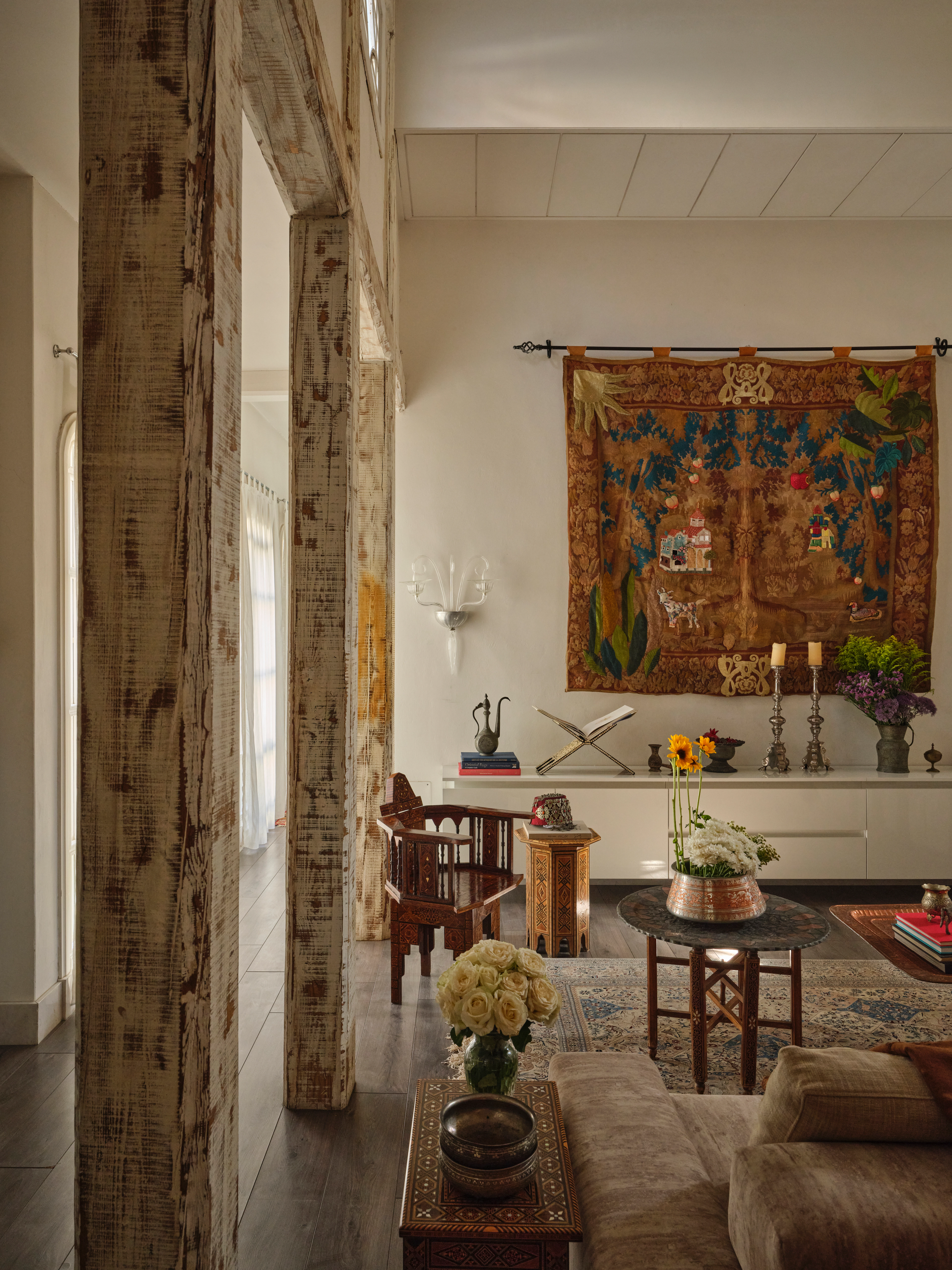 Warm living room with a large woven wall hanging, carved wooden furniture, patterned rug, and floral arrangements.