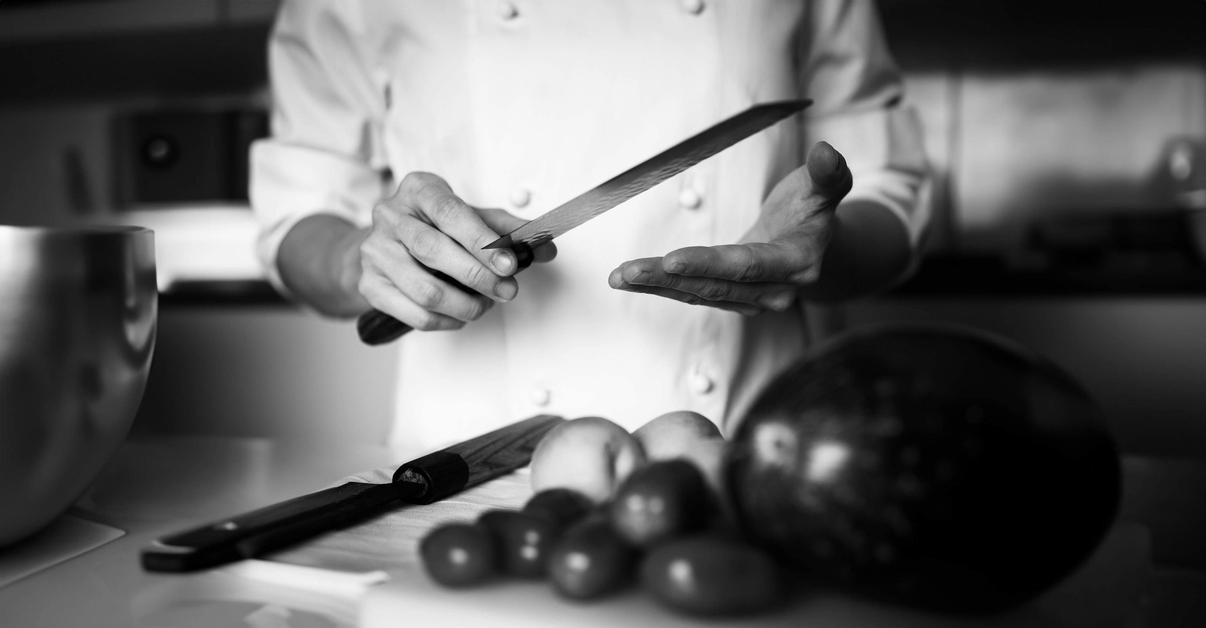 Foto in bianco e nero di una persona vestita da chef. Ha le mani sollevate e impugna un coltello. Inquadratura molto vicina.