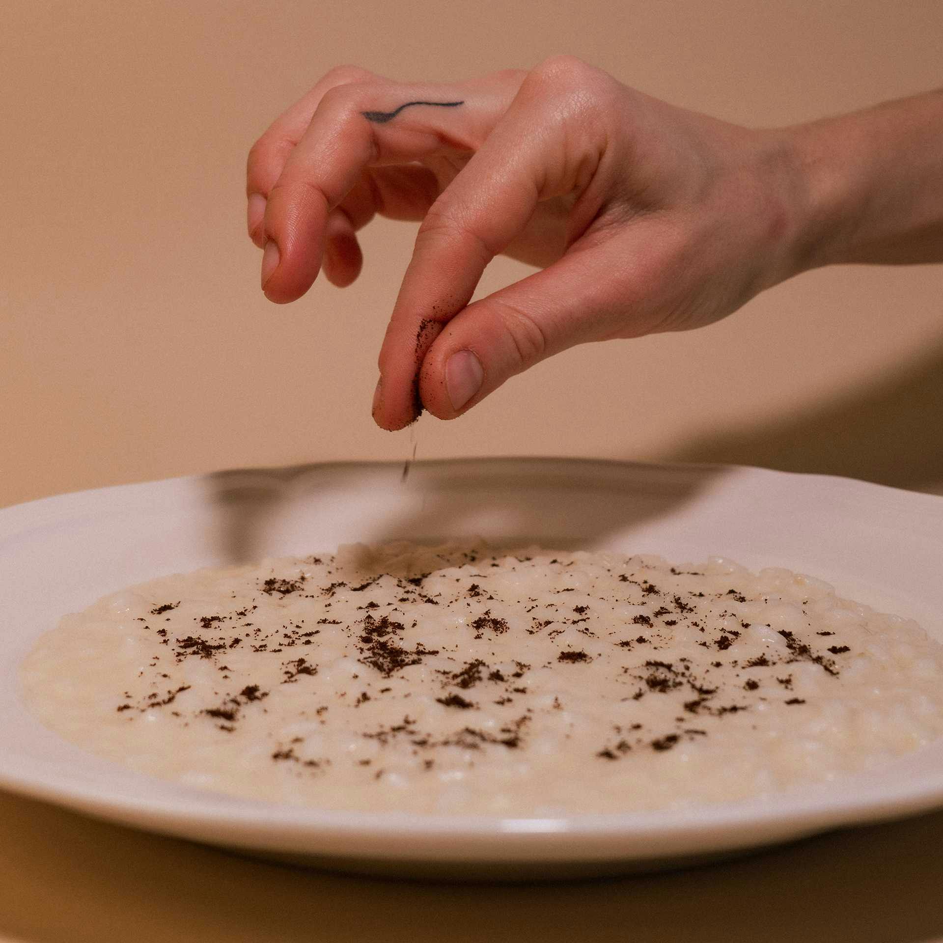 Una mano che spolvera un pizzico di polvere scura sopra un risotto bianco in un piatto di ceramica bianco, creando un'ombra
