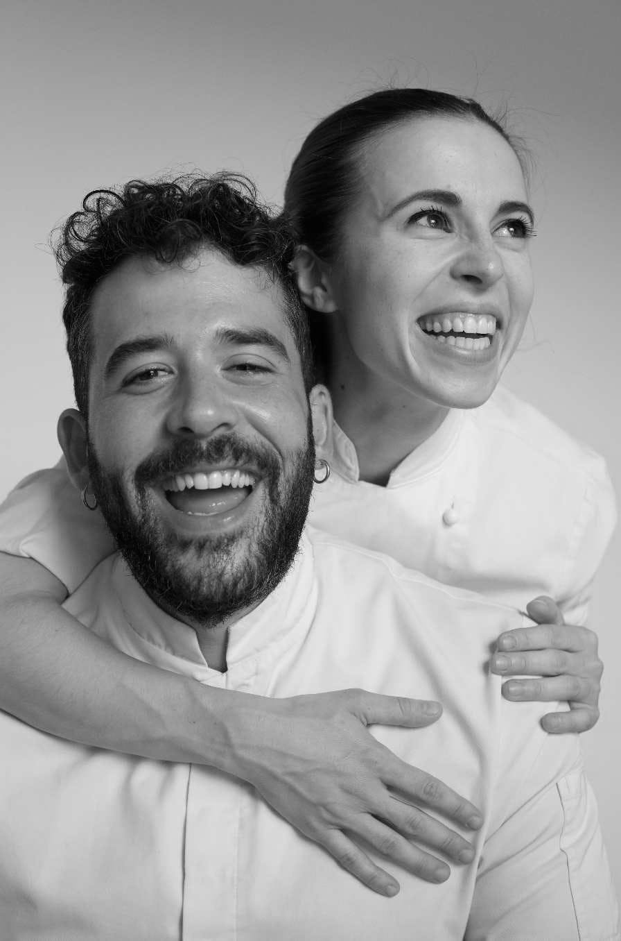 Gabriel Cinque e Valeria Loi, chef e founder di Impronta, in giacca da chef bianca, ridono con gioia, in bianco e nero.