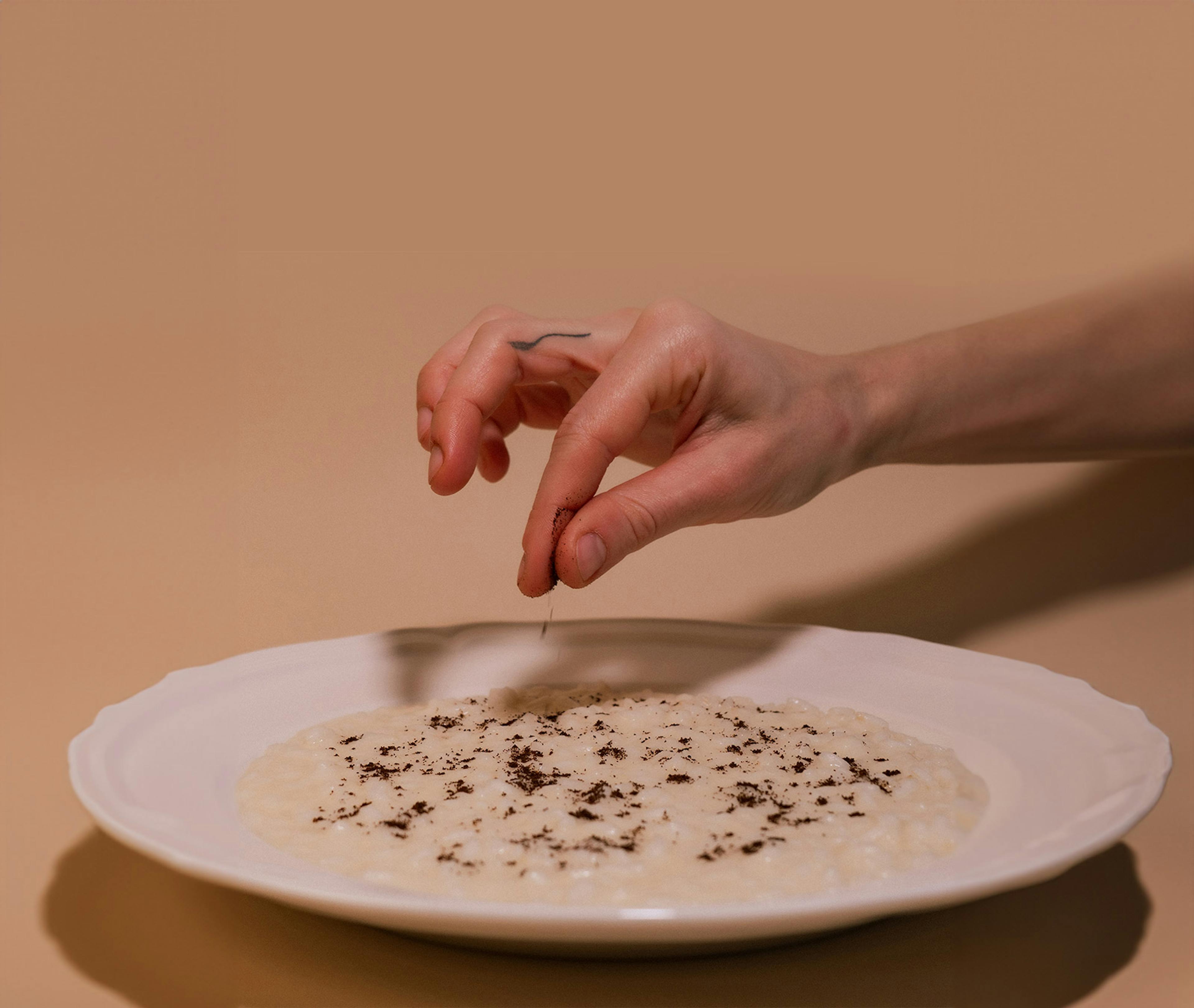 Una mano che spolvera un pizzico di polvere scura sopra un risotto bianco in un piatto di ceramica bianco, creando un'ombra.