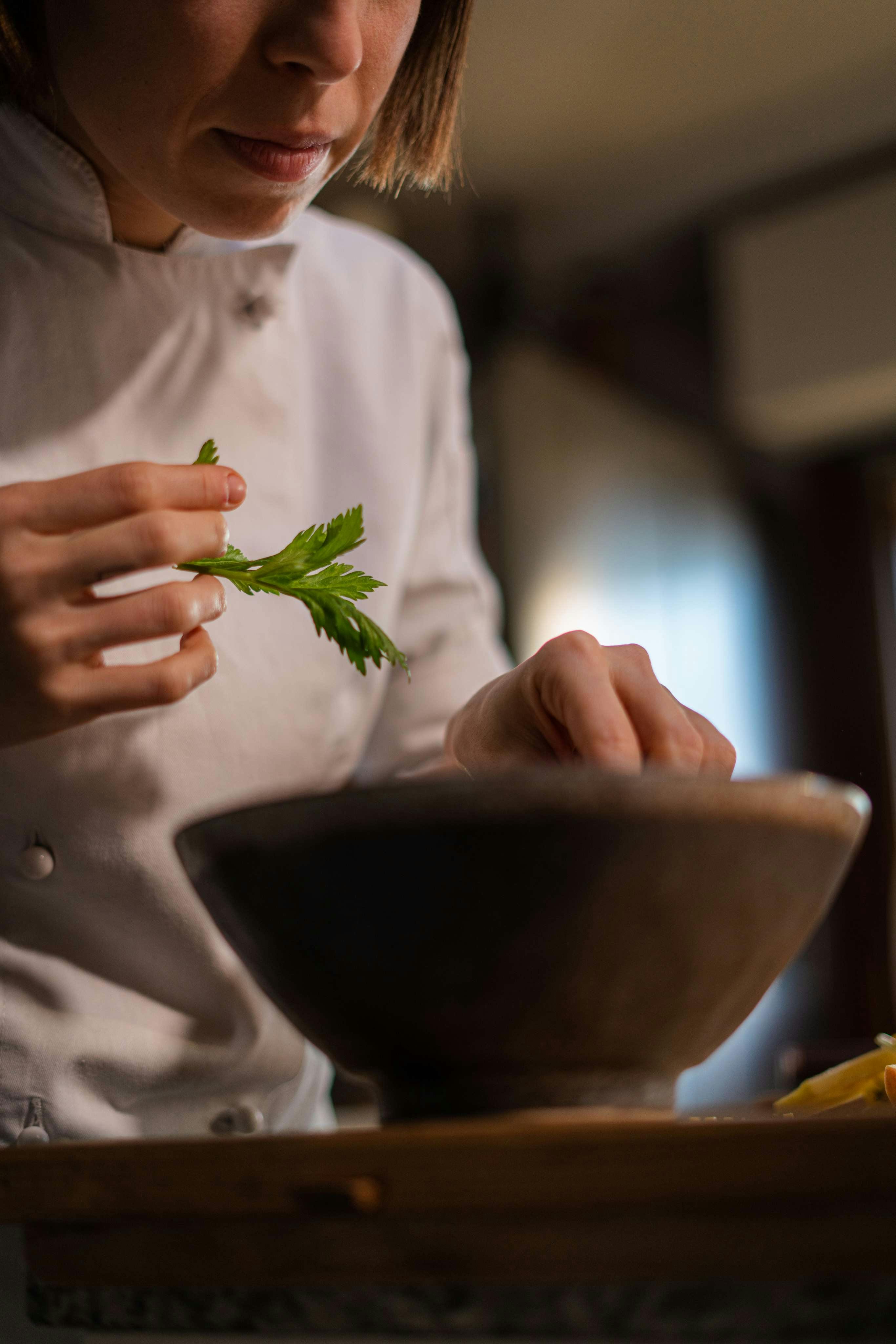 Persona vestita in divisa da chef (giacca bianca) che sta guarnendo un piatto con delle foglie di levistico (verde acceso).