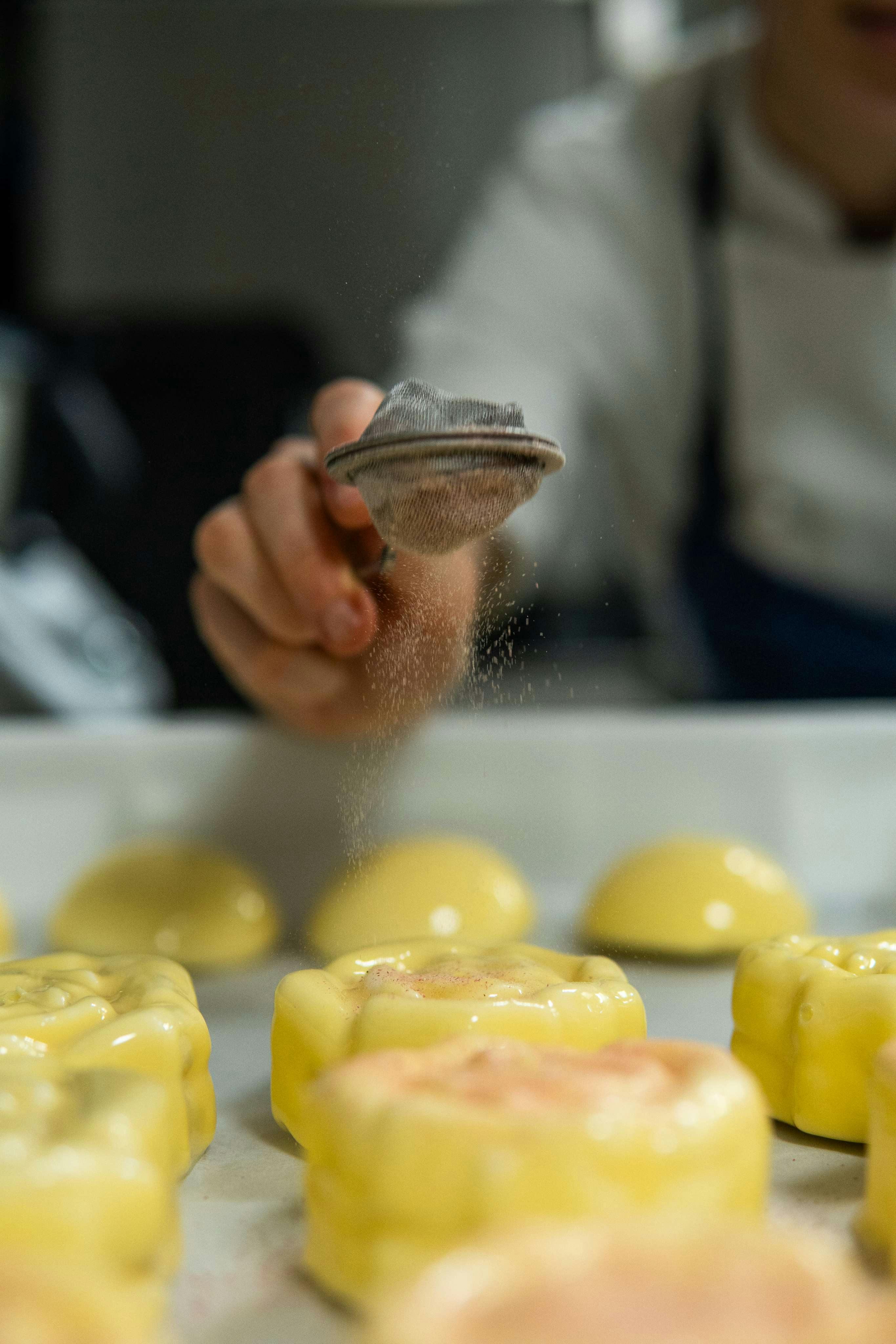 Dolci glassati di colore giallo crema; una mano tiene un colino da cui scende una polvere rosa di rabarbaro essiccato.