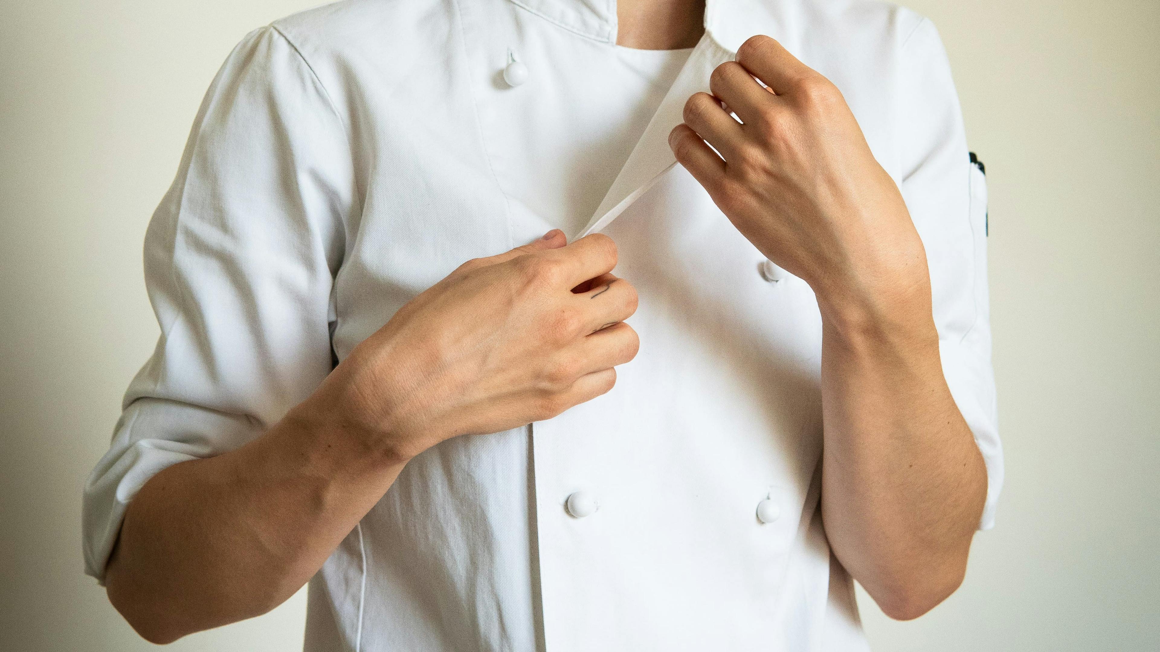 Una persona vestita con la divisa da chef (giacca bianca doppiopetto), nell'atto di allacciare la giacca stessa.