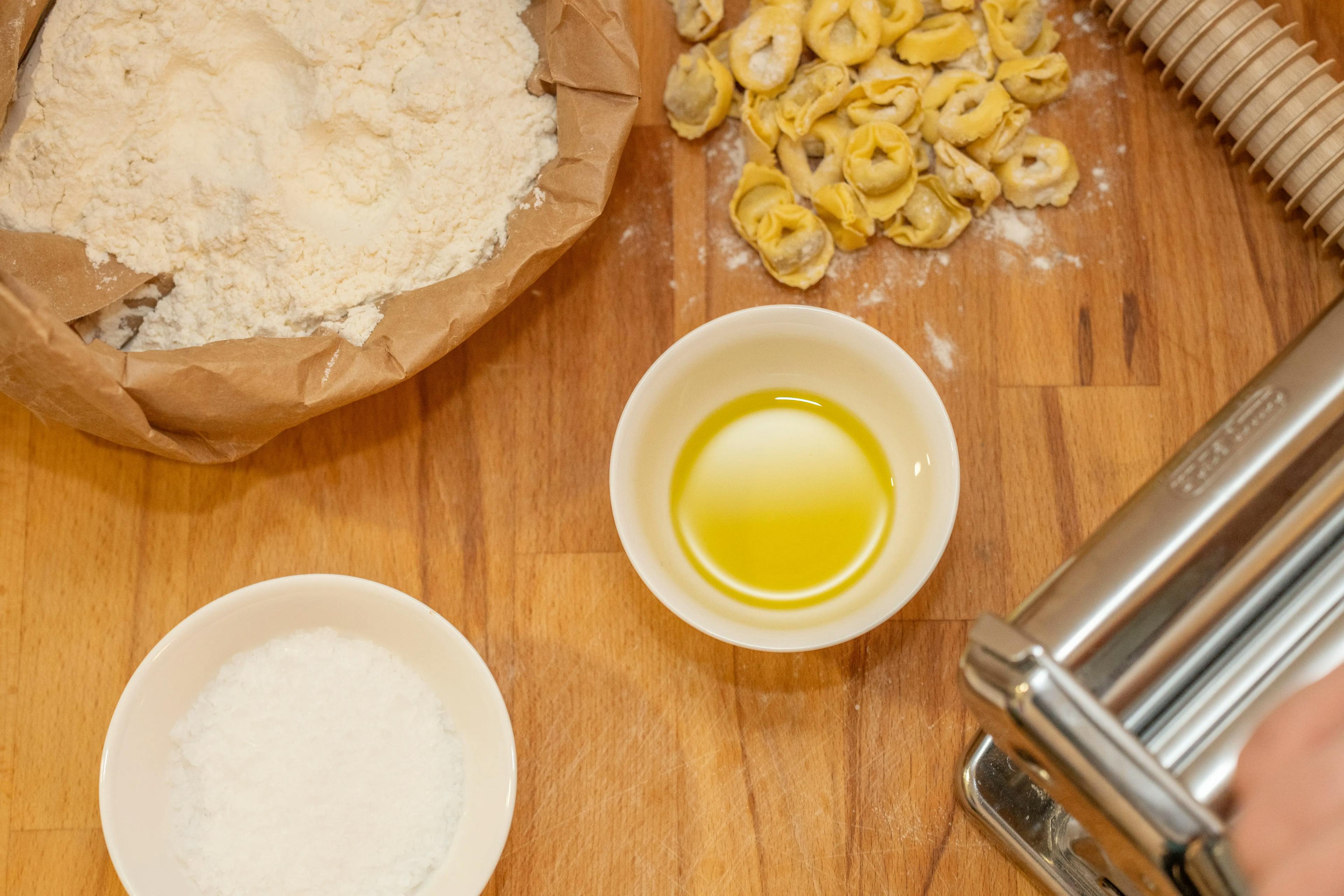 Posizionati su un ripiano di legno: ciotolina di olio, sacchetto di farina, tortellini, macchina per stendere la pasta.