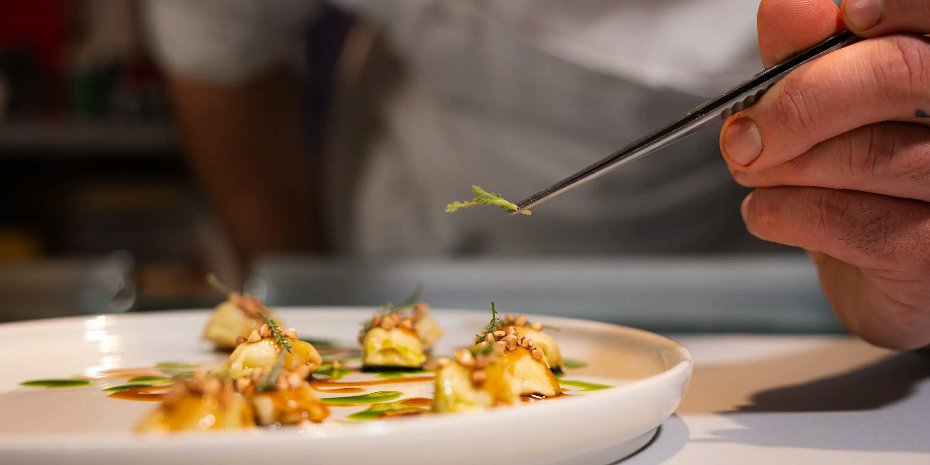 Fogliolina di erba aromatica tenuta da una pinzetta di precisione per guarnire un piatto di pasta fresca.