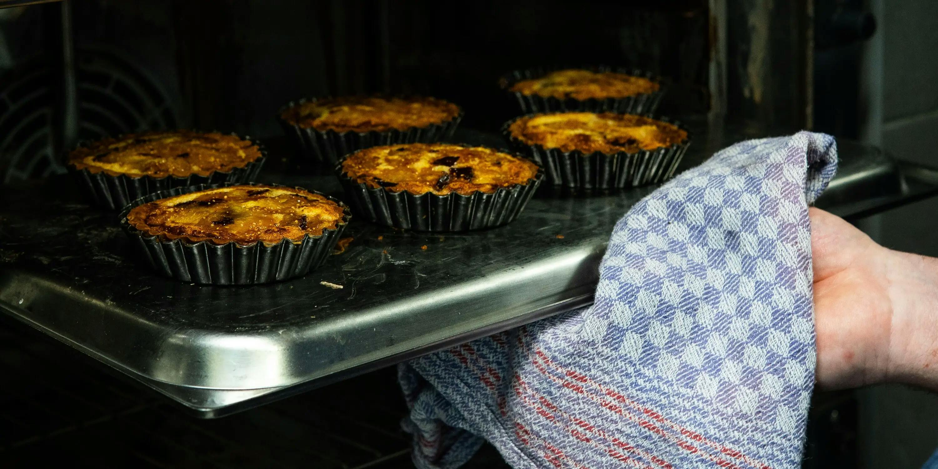 Una teglia da forno, con sopra delle tortine, impugnata da una mano con uno strofinaccio da cucina a quadri blu.