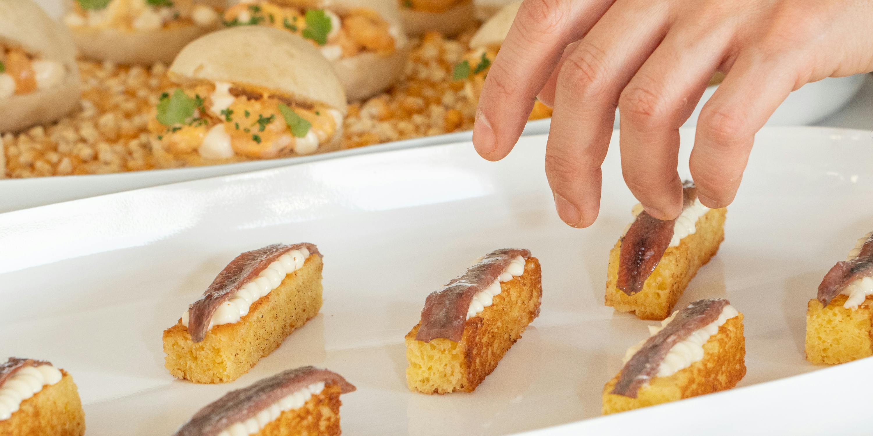 Buffet di finger food (pan brioche, burro, acciughe; mini bao con gamberi) con la mano di un cliente che sta per servirsi.