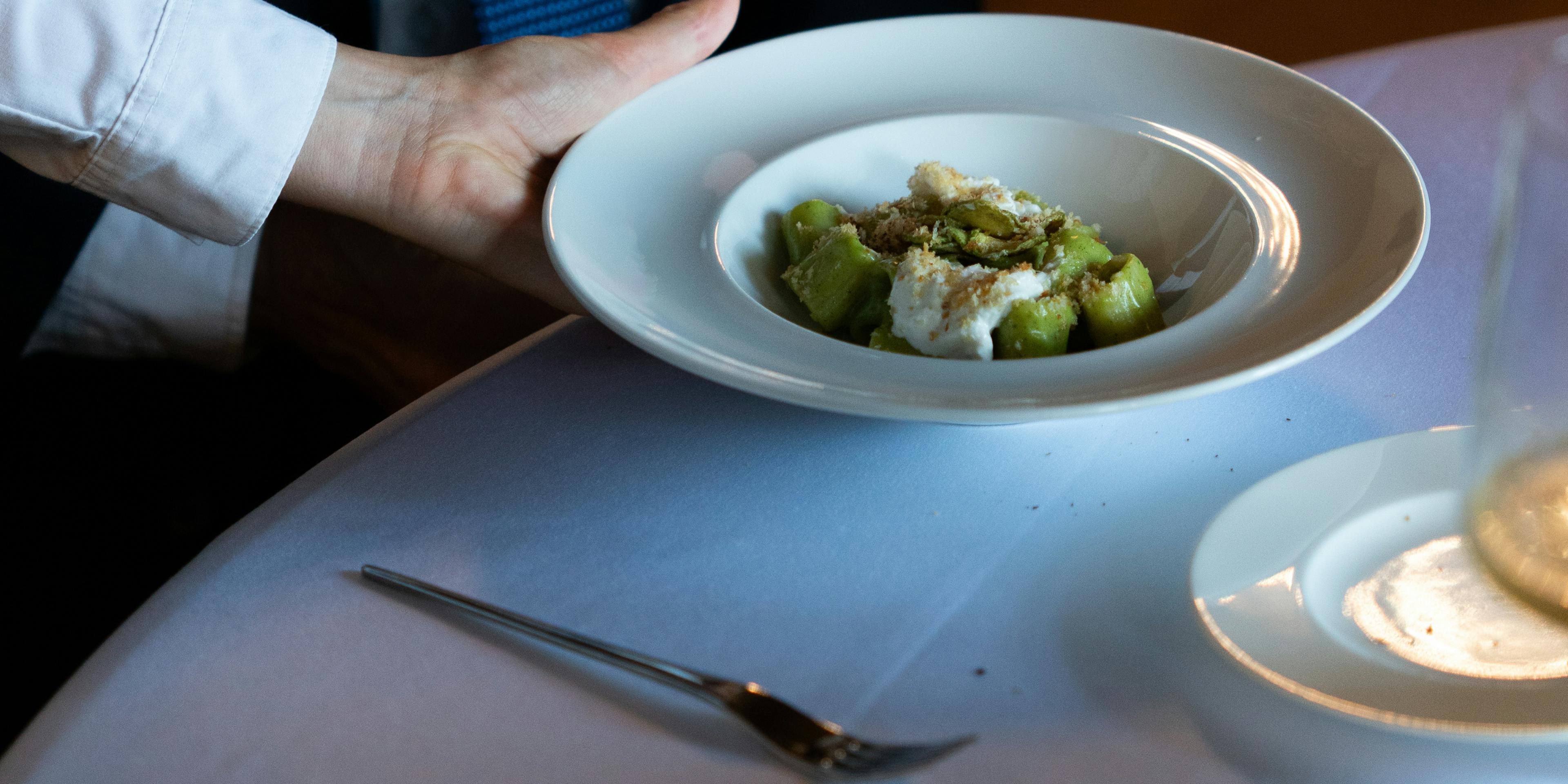 Un piatto di pasta di Impronta viene servito con eleganza a un cliente, su tovaglia bianca. Inquadratura molto ravvicinata.