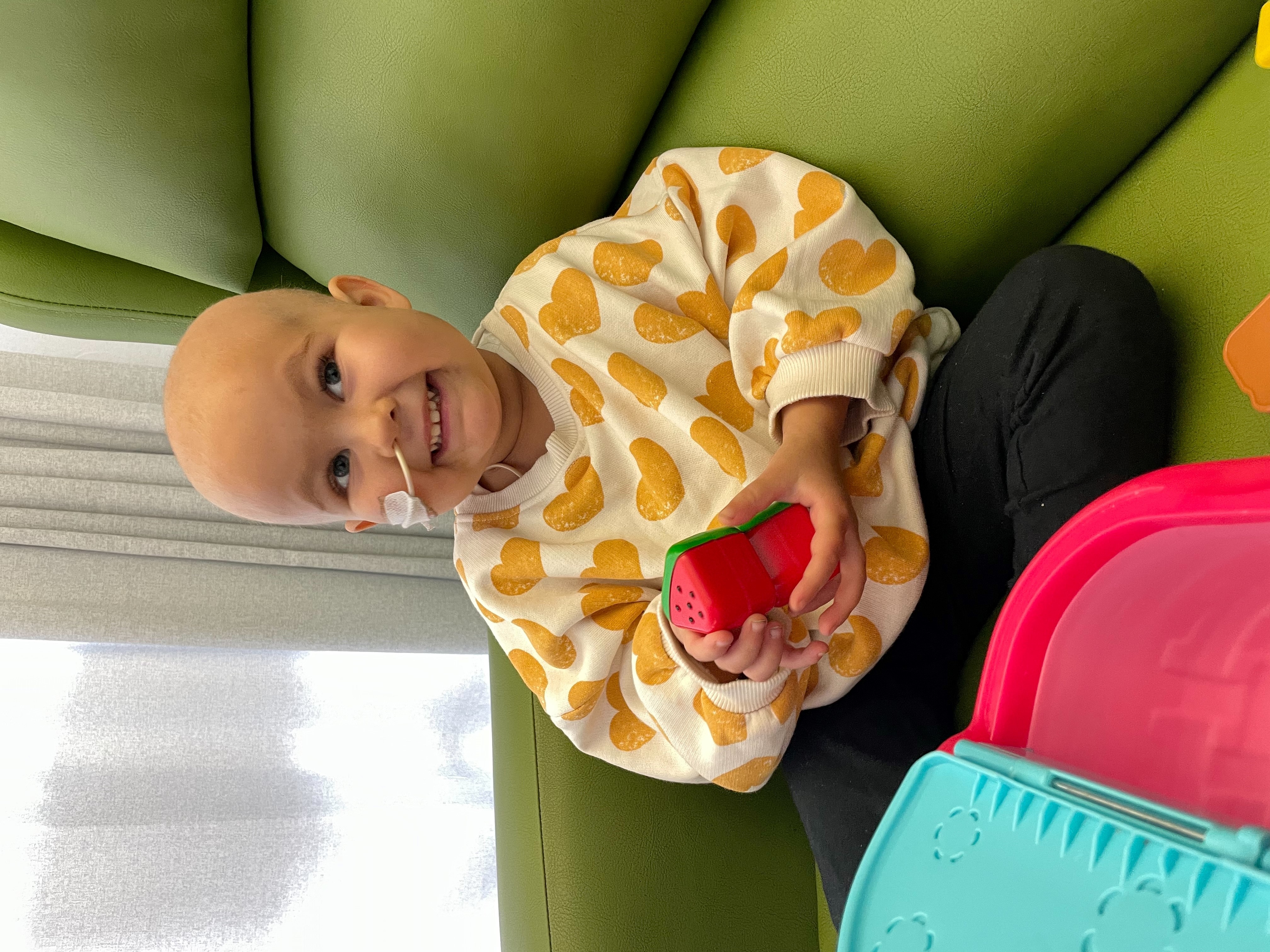 Picture of Cora sitting upright on a bright green chair. She is wearing a white jumper with yellow hearts. She is holding a toy. 