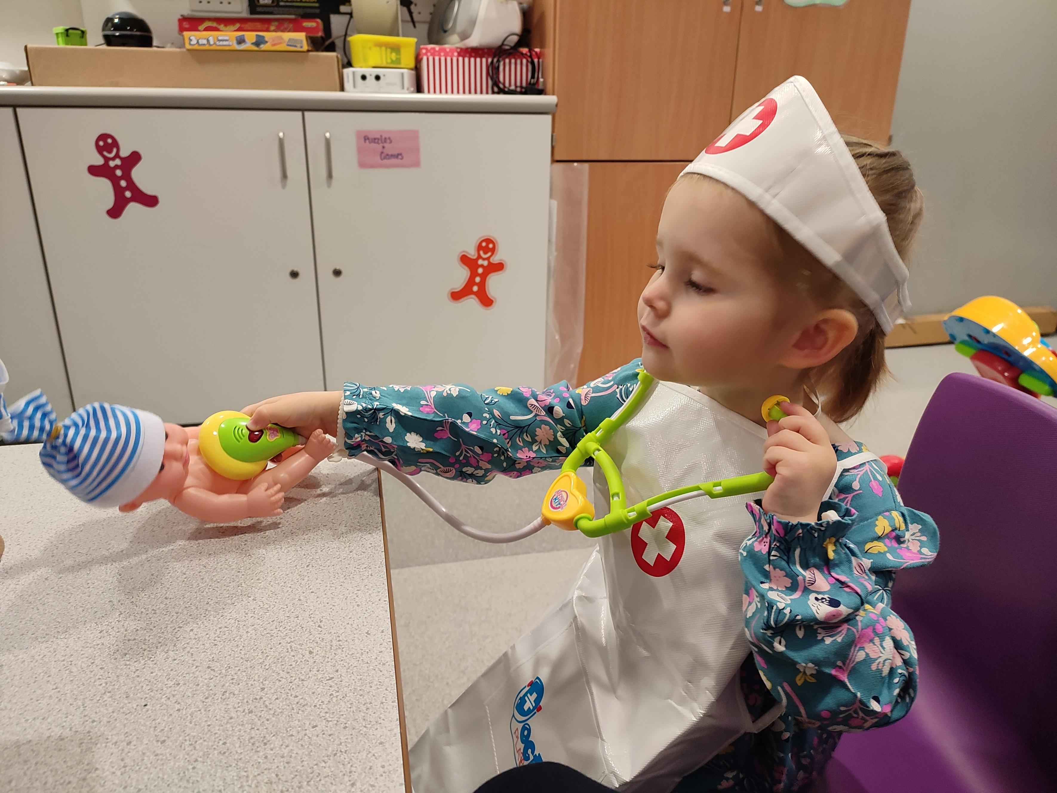 Fiony dressed as a nurse, sat at a table, using a toy stethoscope to listen to heart of a doll