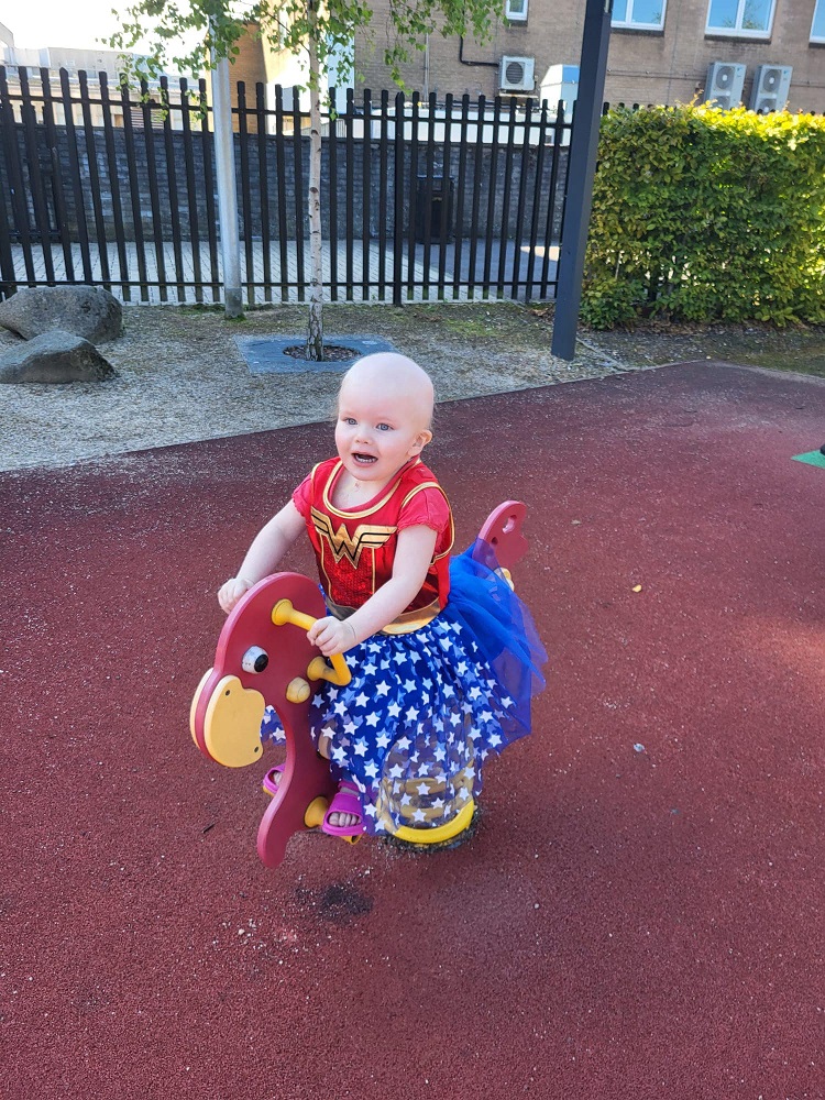 Indy is at a playpark, riding on a play horse. She is wearing a 'Super Woman' costume. 