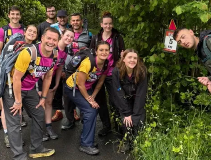 The team posing during the Gower Peninsula Challenge, stood in front of a sign with greenery in the background.