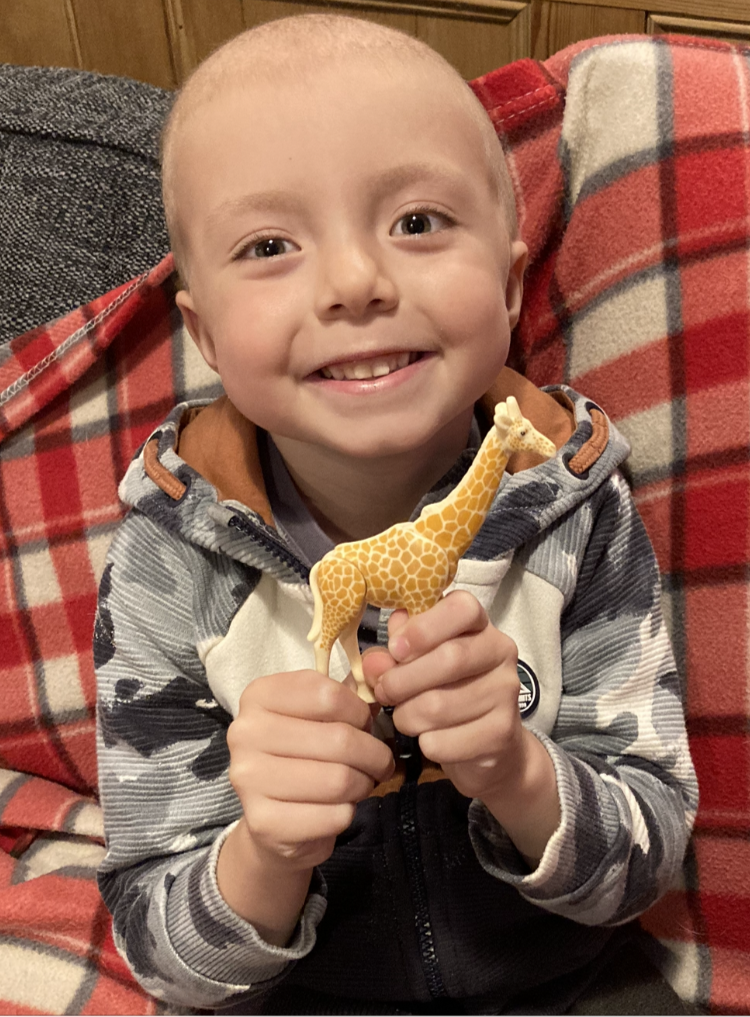 Ollie sitting on a blanket smiling and looking at the camera and holding a small toy giraffe. 