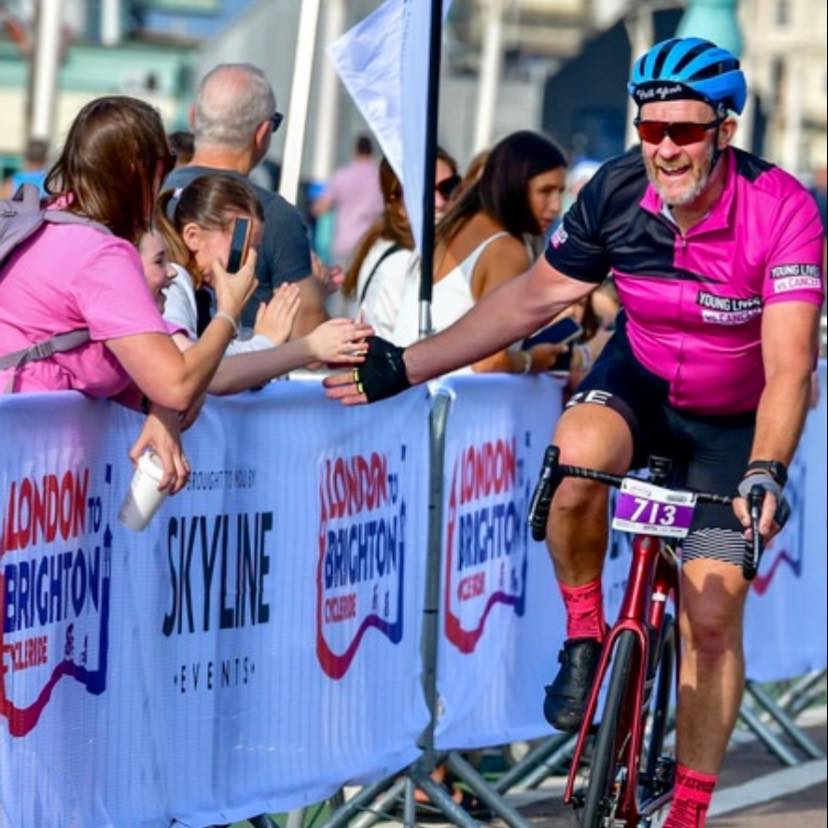 Olivias dad on a bike, high fiving a supporter, wearing Young Lives vs Cancer branded cycle gear