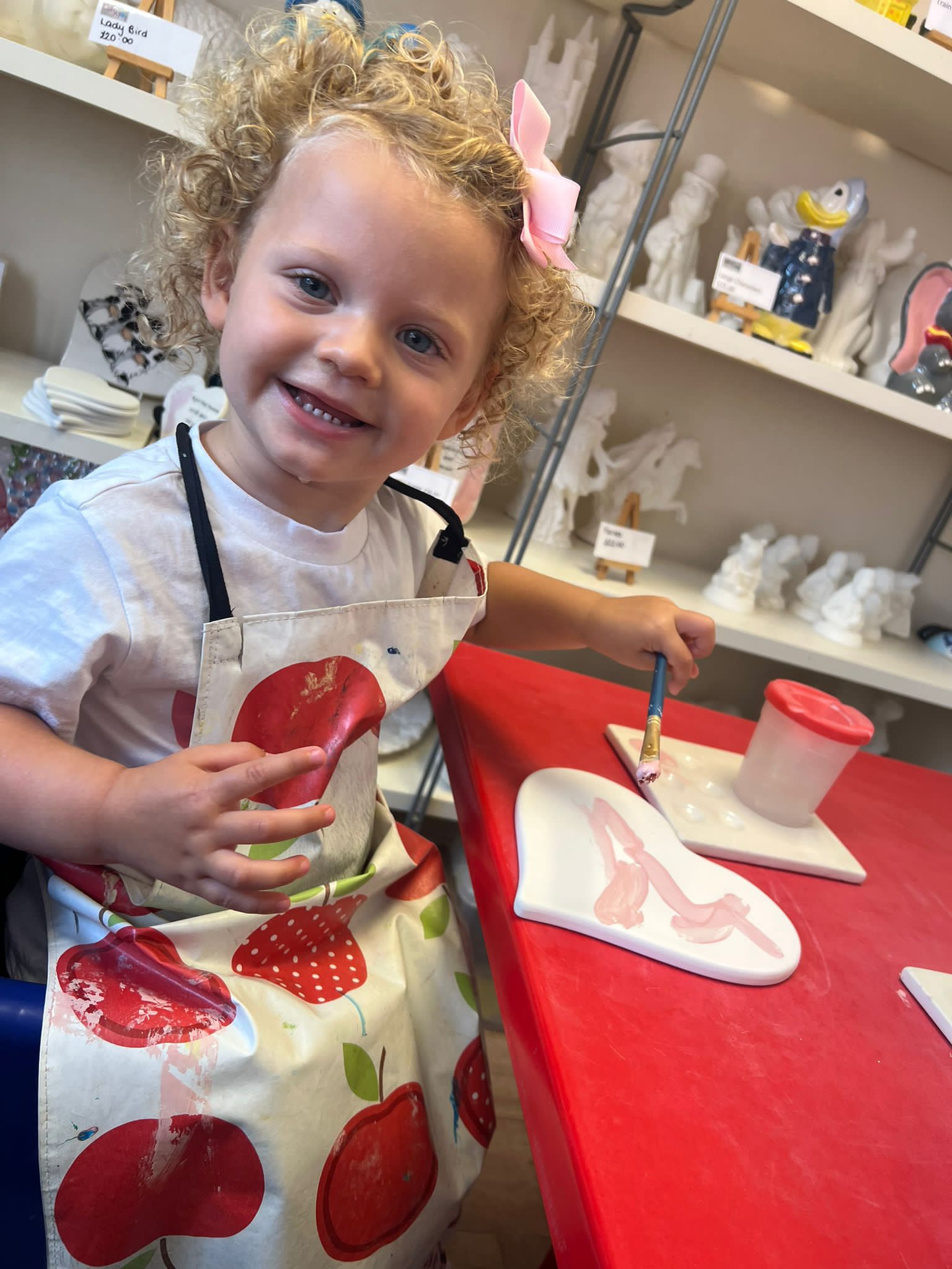 ivy sat at a table in an apron, painting, with a pink ribbon in her hair