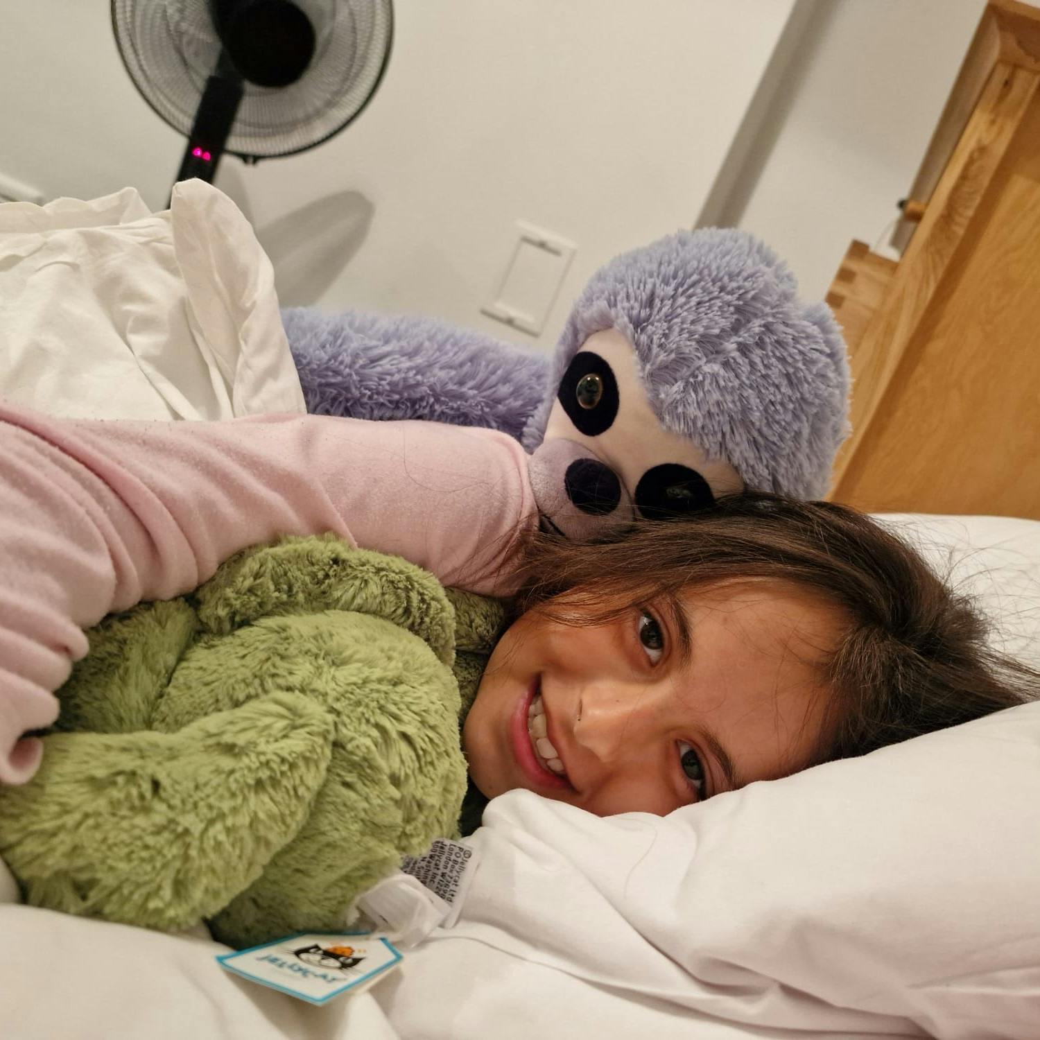 Xanthia lying on her side on a bed hugging a green cuddly toy and with a soft toy sloth behind her. She is smiling at the camera