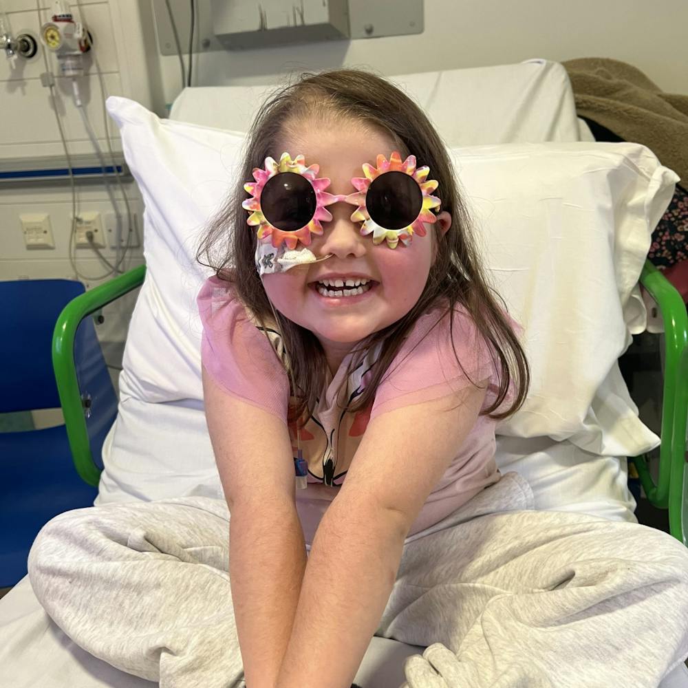 Arabella is on the hospital bed, smiling with funny sunglasses on. She is wearing a pink shirt and squeezing her arms.