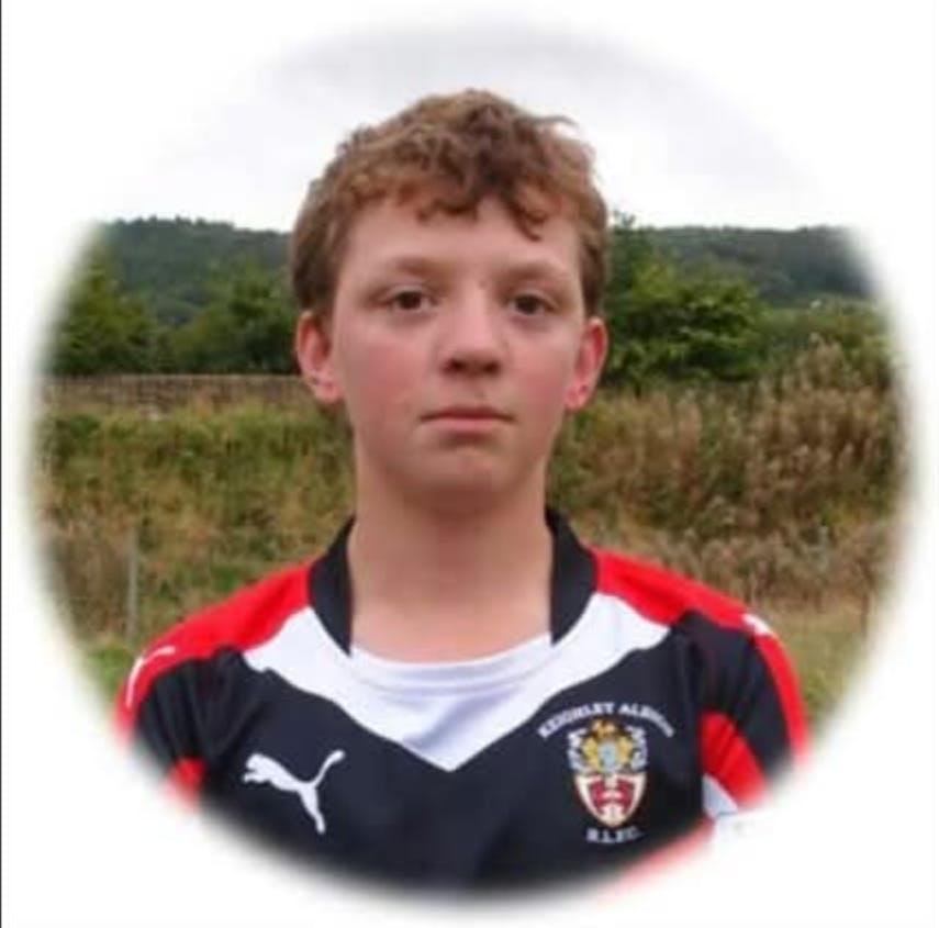 CJ Mynott standing facing the camera, with short curly brown hair. He is wearing a black, red, and white rugby jersey with the logo of Keighley Albion R.L.F.C. He is standing outdoors in front of greenery and hills, looking directly at the camera with a neutral expression. The image has a soft white vignette border.