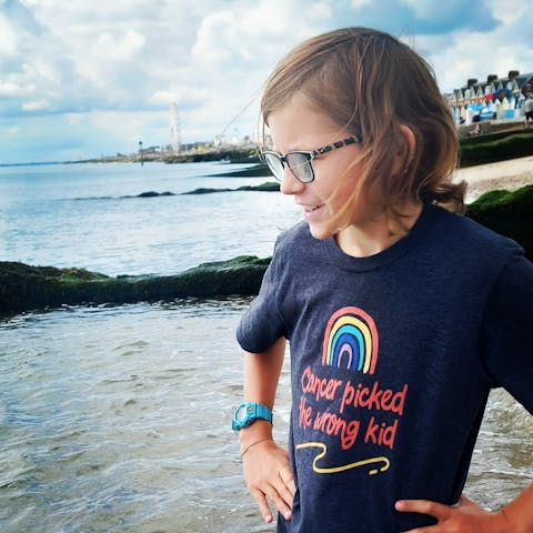 Remus is standing by the water of the seas edge with his hands on his hips. He is wearing a dark navy t-shirt that has a rainbow on the front saying 'cancer picked the wrong kid'
On his right wrist he is wearing a blue watch. he is wearing black glasses and the wind is blowing his hair.