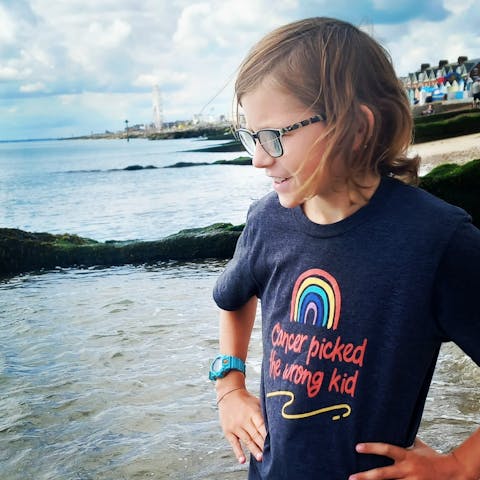 Remus is standing by the water of the seas edge with his hands on his hips. He is wearing a dark navy t-shirt that has a rainbow on the front saying 'cancer picked the wrong kid'
On his right wrist he is wearing a blue watch. he is wearing black glasses and the wind is blowing his hair.