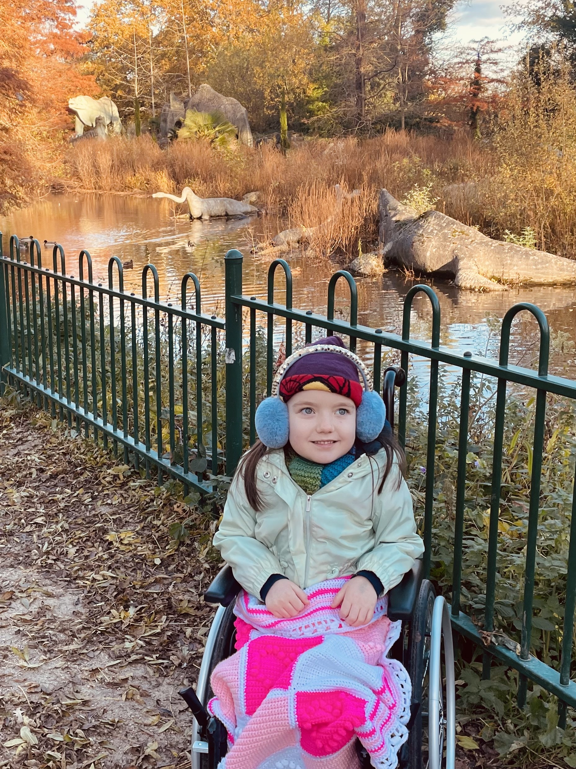 Ava is sitting in her wheelchair in a park. In the background there is a small river protected by a black metal fence, and on the other side of the river there are  three dinosaur statues. It's a bright winter day and Ava is wearing a pink and white blanket on her legs, a green jacket, purple beanie hat and a pair of light blue earmuffs. 