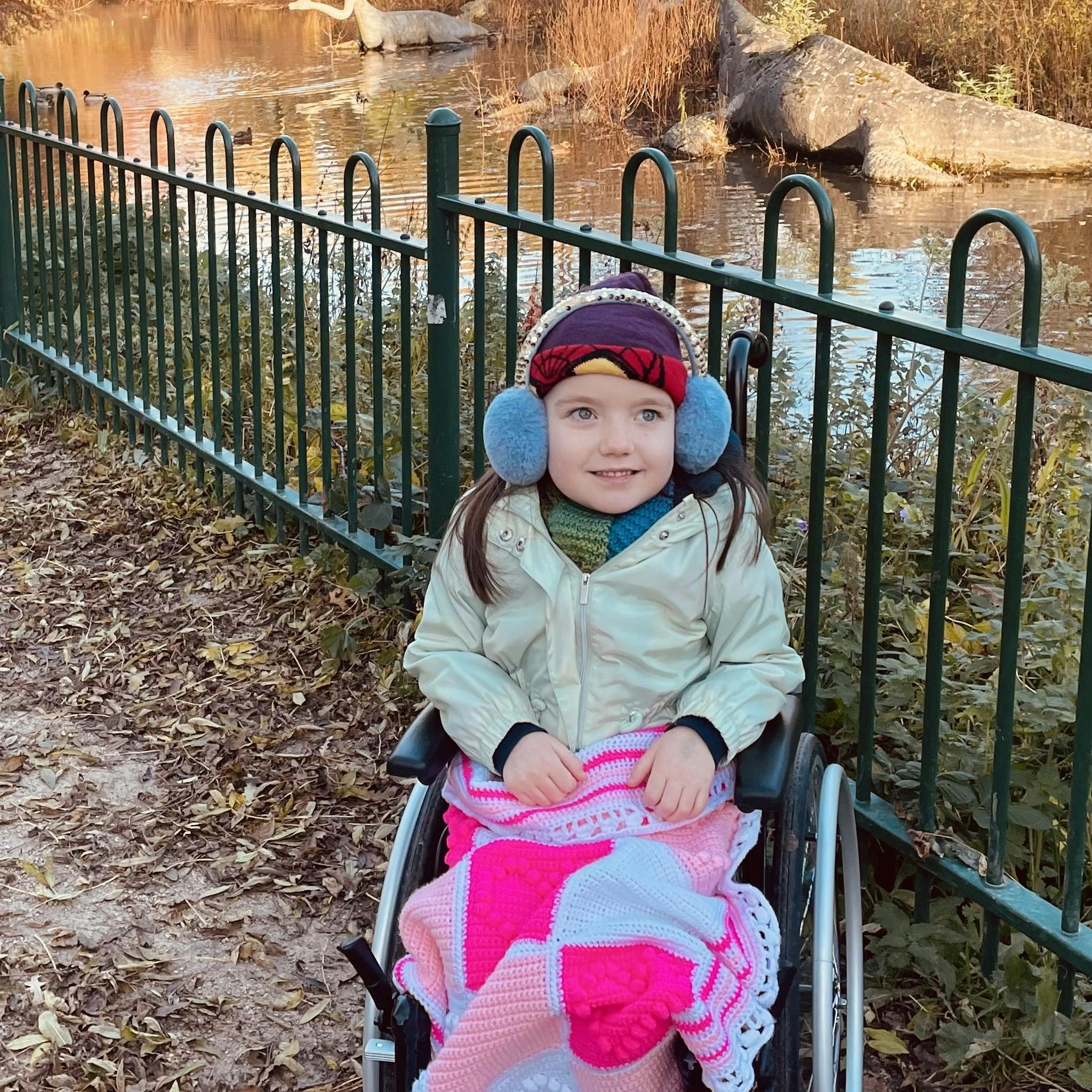Ava sat in a wheelchair with a pink checked blanket over her lap, wearing blue ear muffs. She is in front of a metal railing with a pond in the back ground