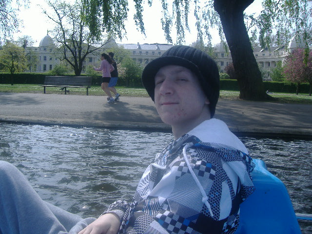 Ryan sat in a boat on a lake in a park, looking at the camera, he has a black hat on and black and white check hoodie