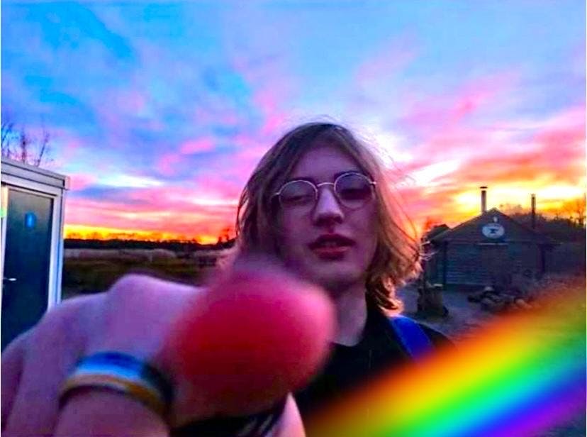 An image of Sidney pointing at the camera, he is wearing a ring with a rainbow on and there is a sunset behind him.