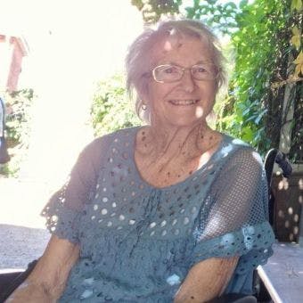 Valerie sitting on a chair, wearing a blue top with frill and holes in, wearing glasses, smiling at te camera