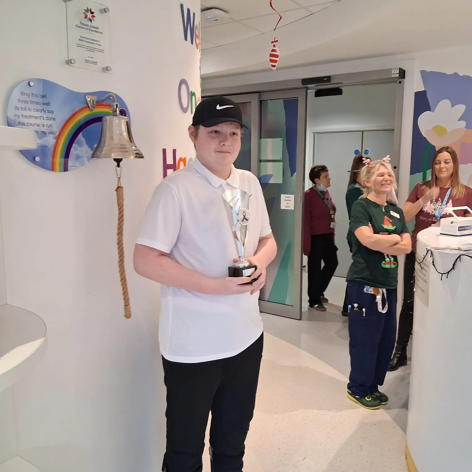 Jakub stands on a hospital ward wearing a white polo shirt and black cap, holding a silver trophy beside a wall-mounted bell rung to mark the completion of cancer treatment, with staff and decorations in the background.