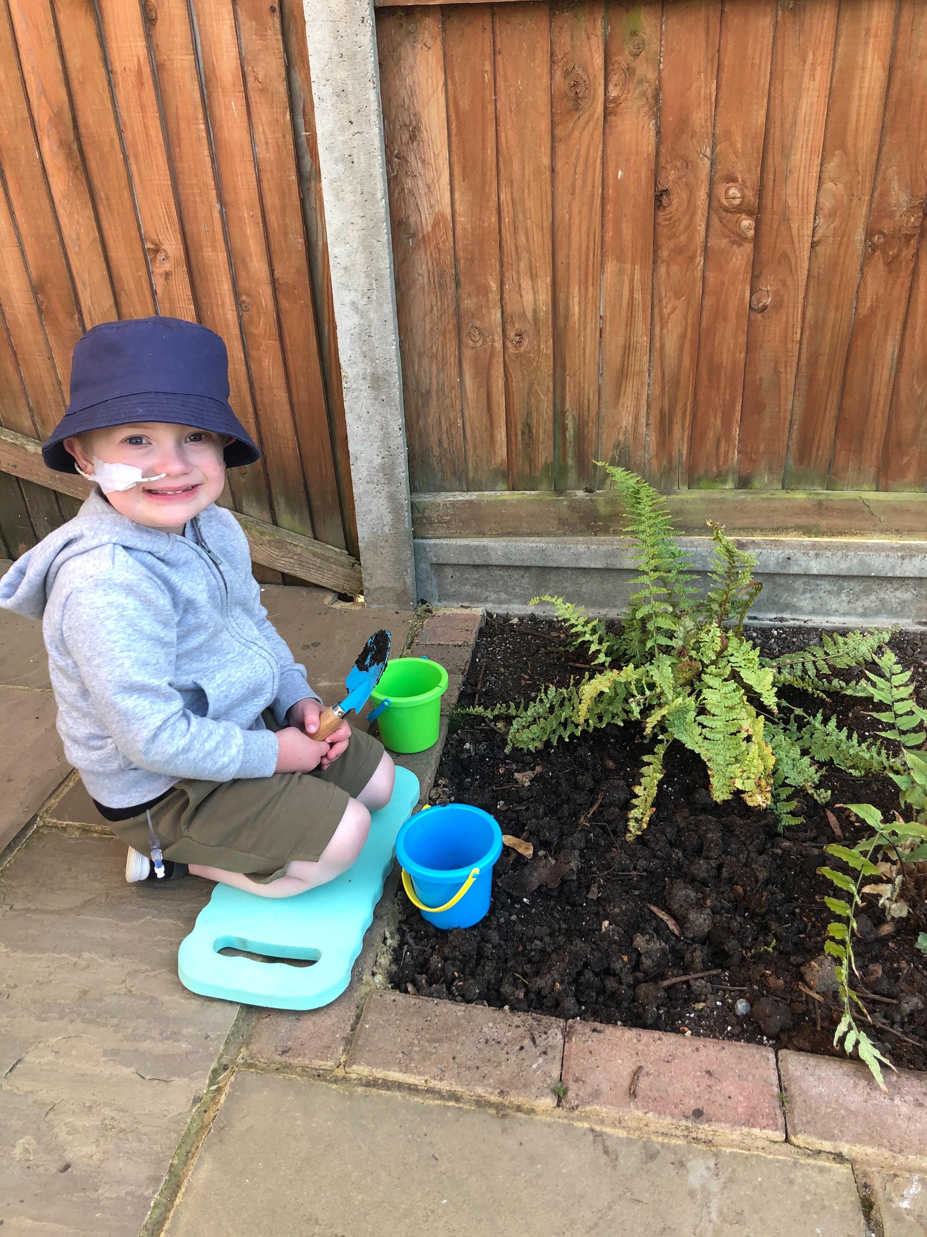 Alfie knelling next to a patch of garden, with a spade in hand, bucket next to him  and a fern in the ground