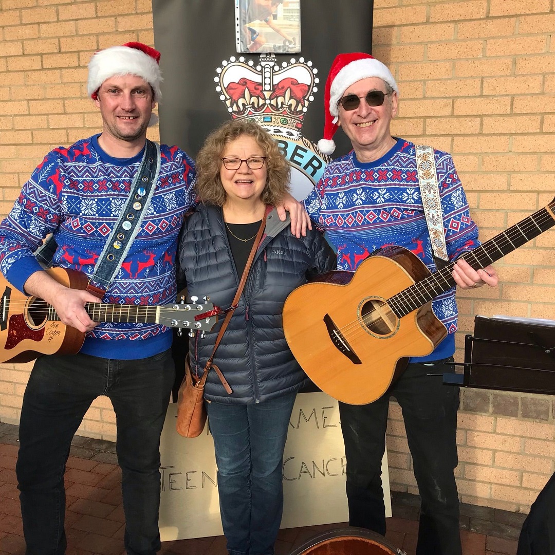 Kevin and Ben holding guitars and wearing christmas jumpers and hats, with Janet between them