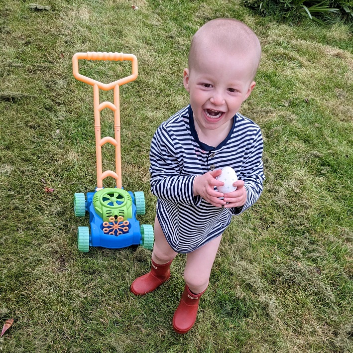 Arthur standing in the garden, smiling, with a stripy t-shirt on, holding an egg with a toy lawn mover next to him