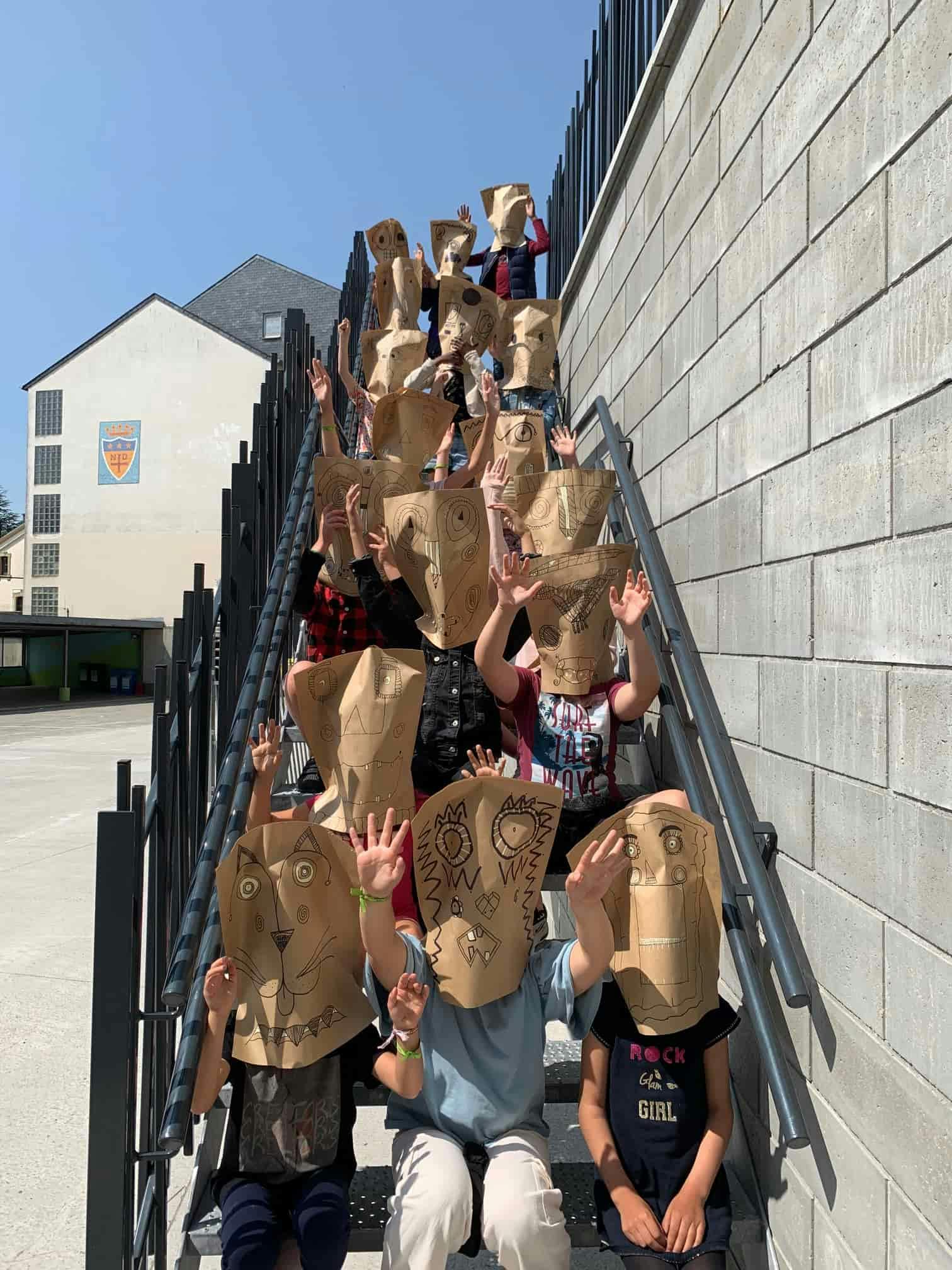 Élèves de l'école à pédagogie active du centre d'Arlon portant des masques en papier et levant les mains.
