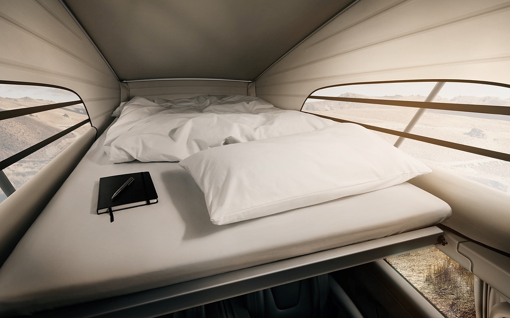 Interior view of a campervan bed with white bedding, pillow, notebook, and pen near a panoramic window.