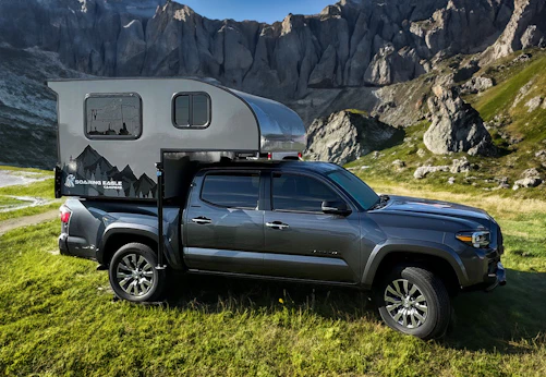 Truck camper parked on green grass with rocky mountains in the background, under daylight.