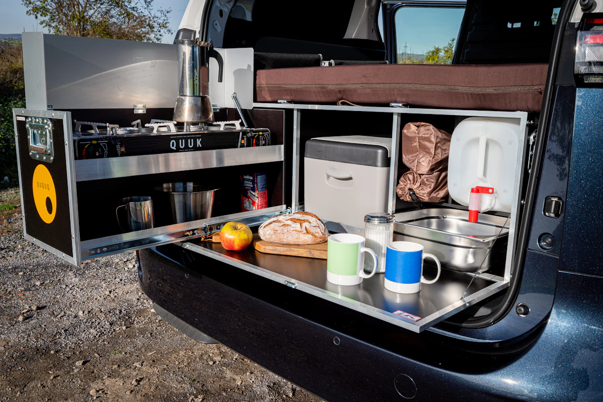 Campervan rear kitchen setup with stove, sink, bread, fruit, mugs, and storage compartments ready for outdoor cooking.