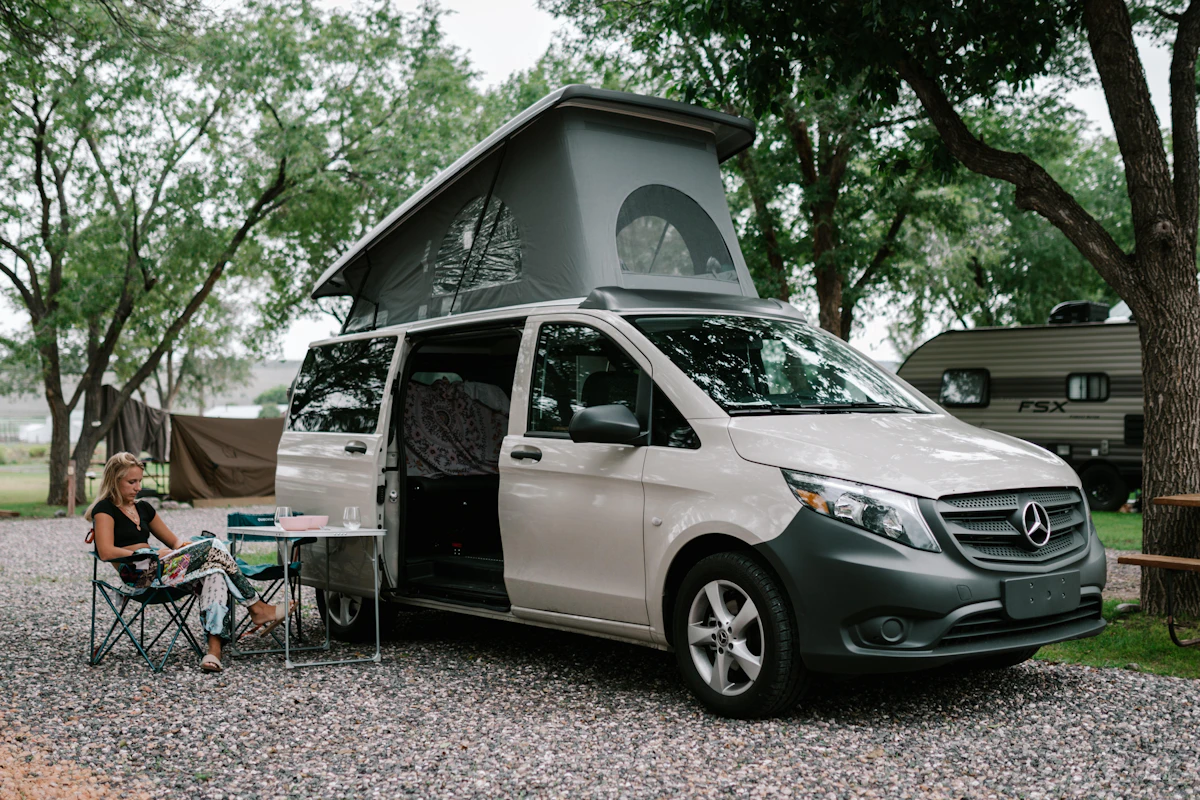 Woman relaxing outside a Mercedes camper van with pop-up roof at a campsite surrounded by trees and other RVs.
