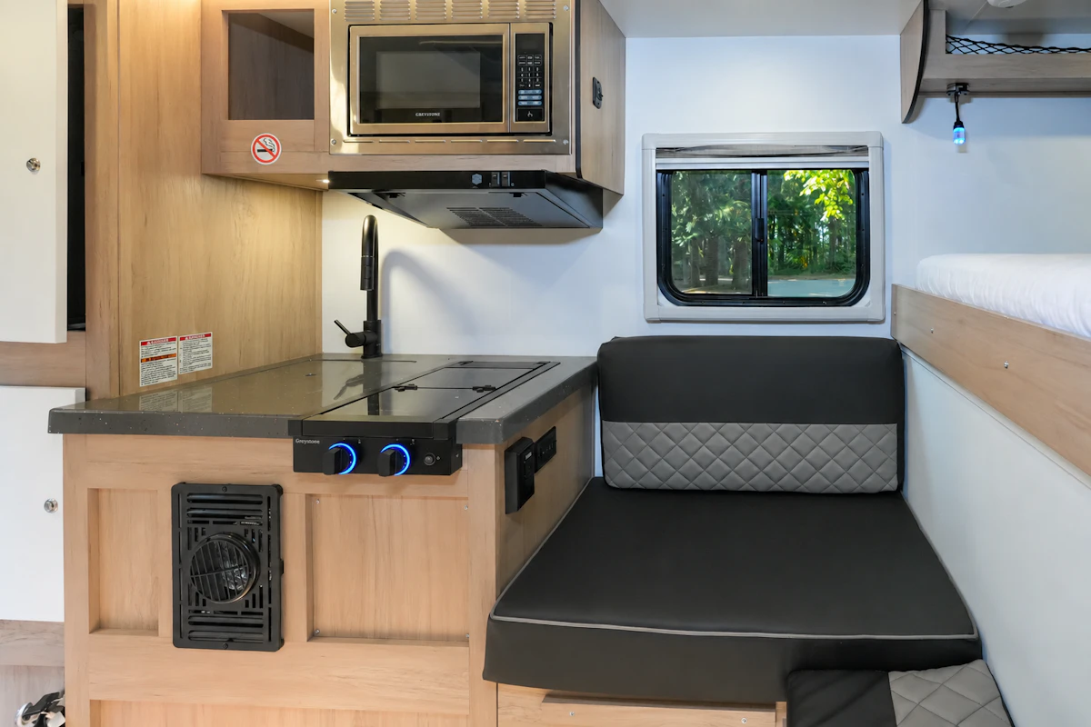 Compact van interior with kitchenette, microwave, sink, stove, small sofa, window, and upper bunk bed visible.