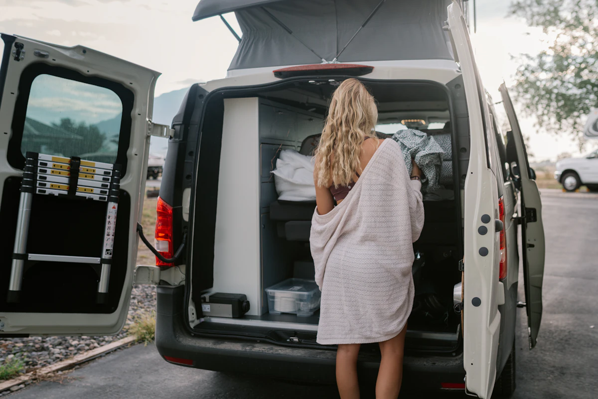 Woman arranging items in the back of an open camper van with ladder attached to the door.
