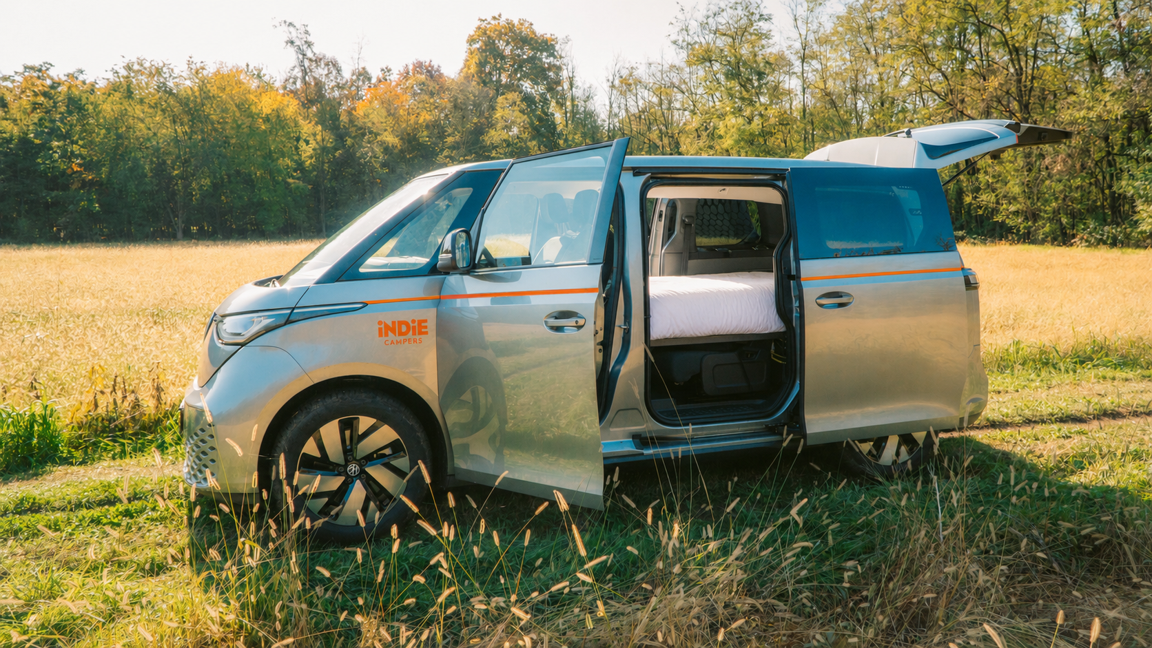 Silver camper van with open doors parked in a field, featuring Indie Campers branding, trees in background.