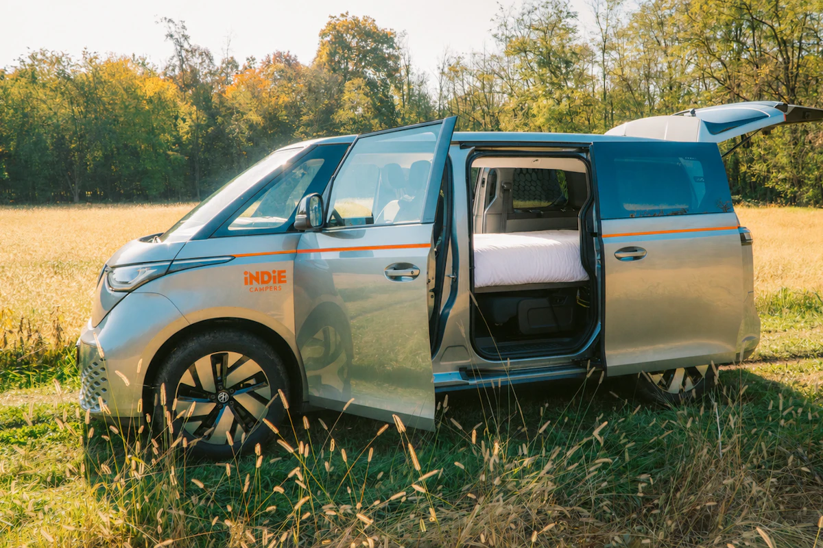 Silver camper van with open doors parked in a field, featuring Indie Campers branding, trees in background.