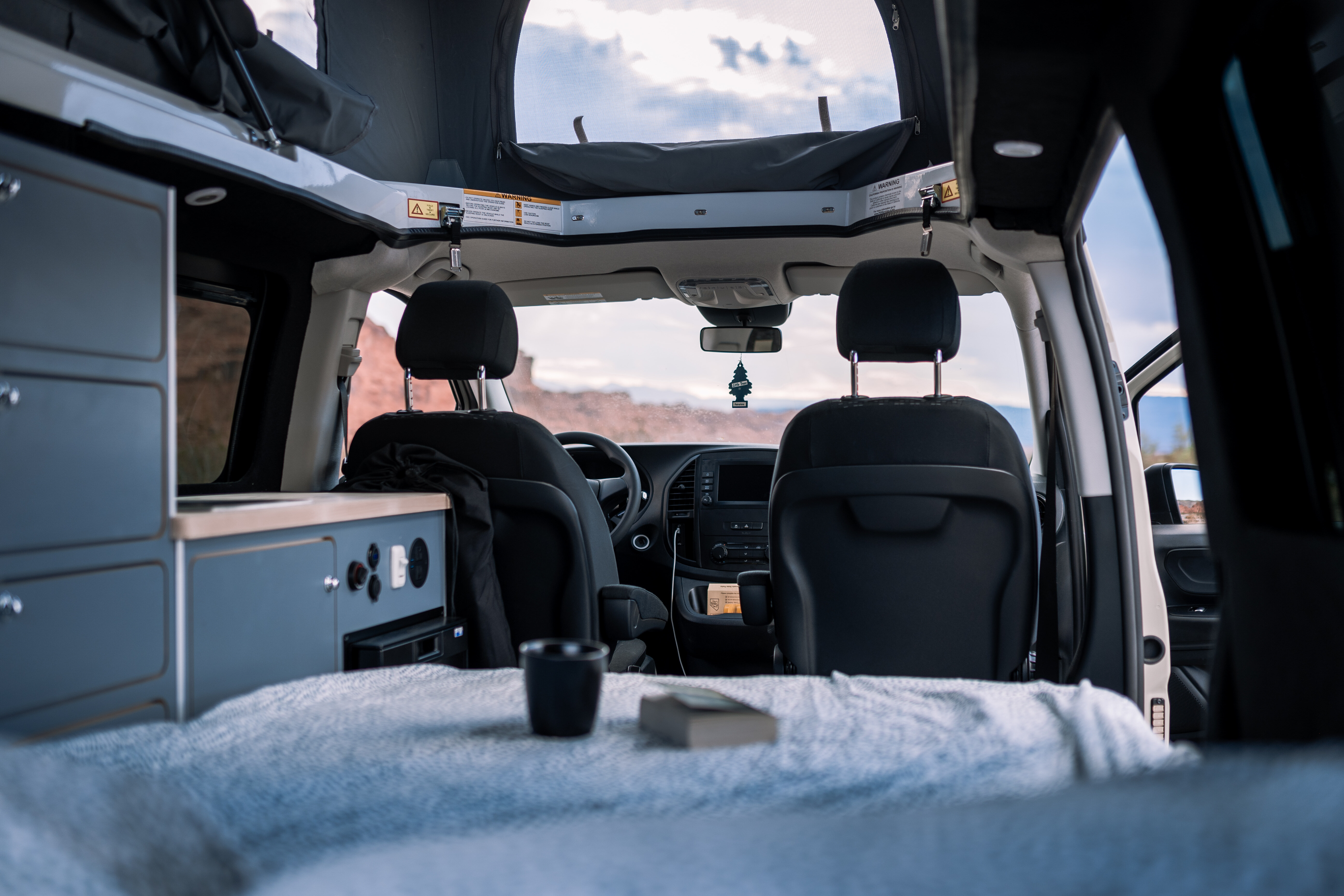 Interior of a campervan with bed, cabinets, front seats, and open roof, parked in a scenic outdoor location.