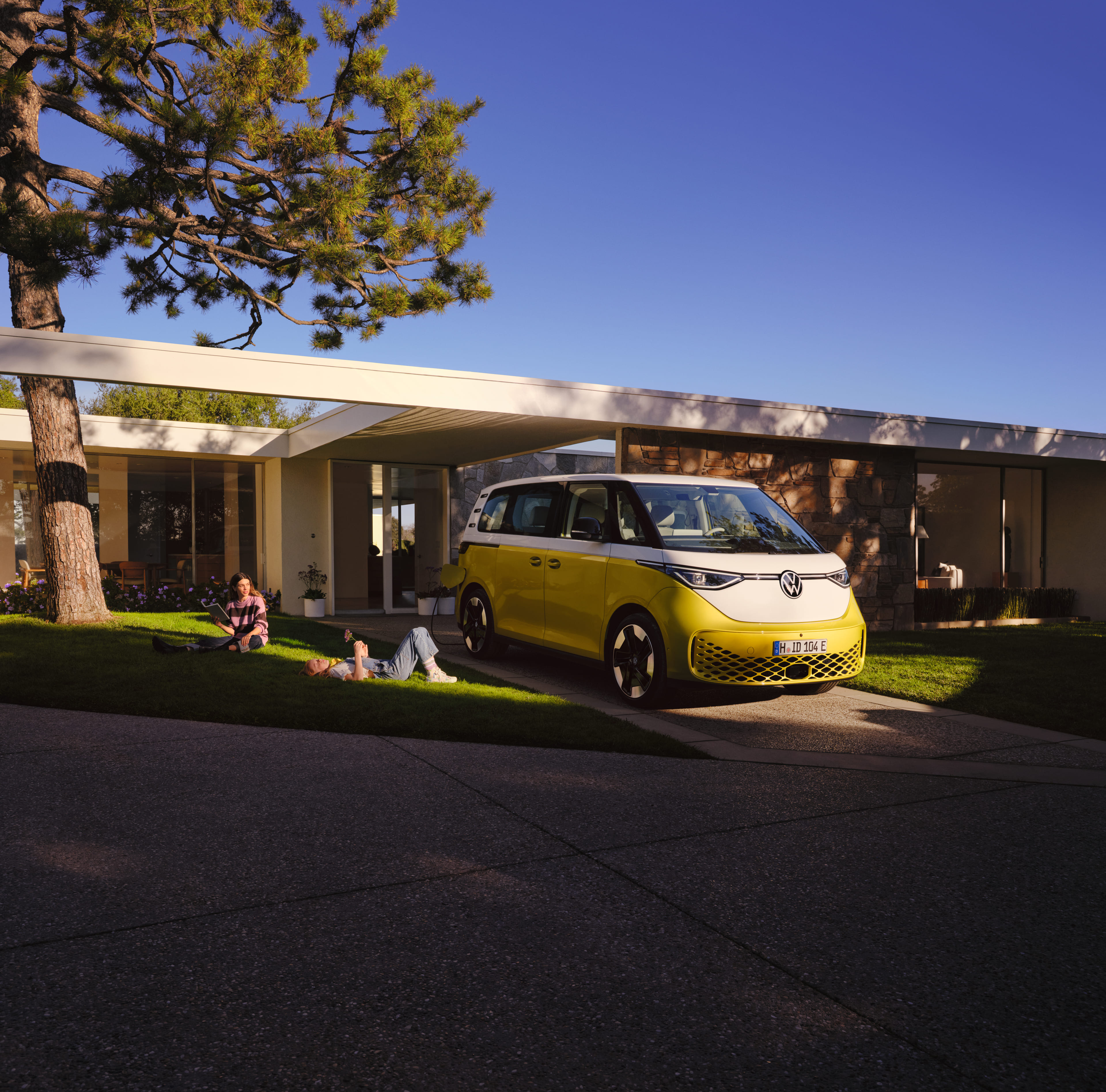 Yellow and white Volkswagen camper van parked in front of a modern house, with people relaxing on the lawn nearby.