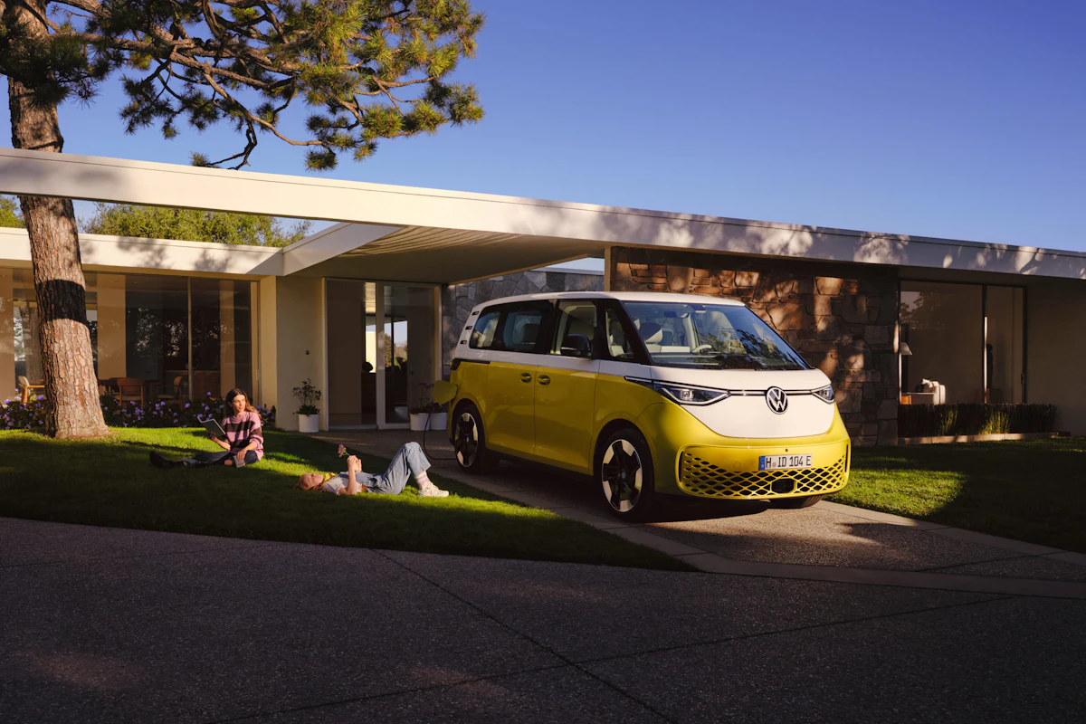 Yellow and white Volkswagen camper van parked in front of a modern house, with people relaxing on the lawn nearby.