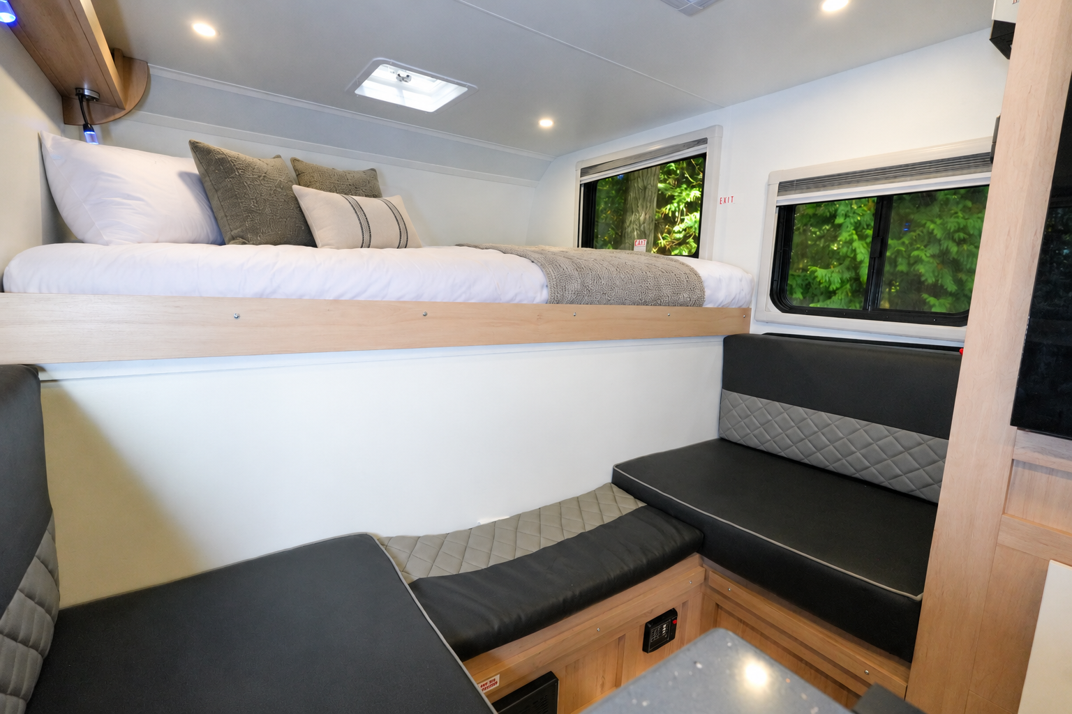 Interior van view with upper bed, lower seating area, overhead storage, windows, and modern lighting visible.