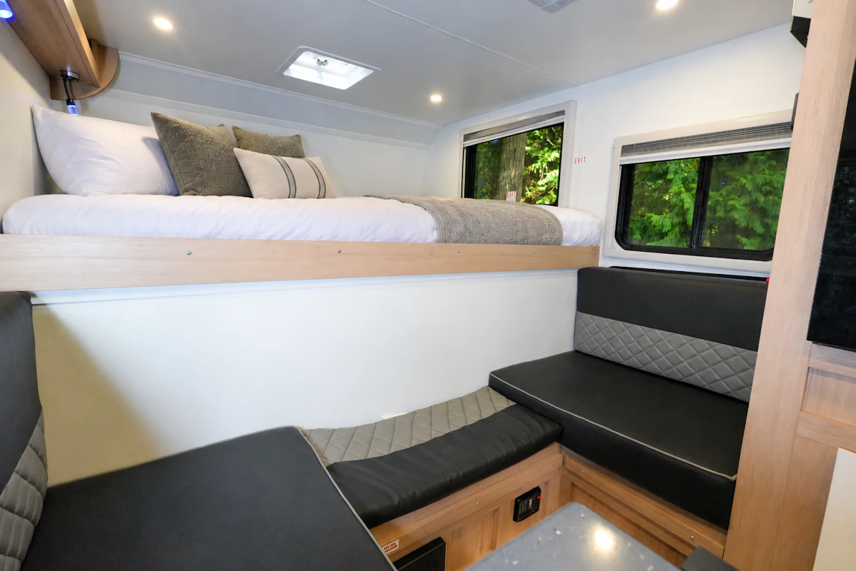 Interior van view with upper bed, lower seating area, overhead storage, windows, and modern lighting visible.